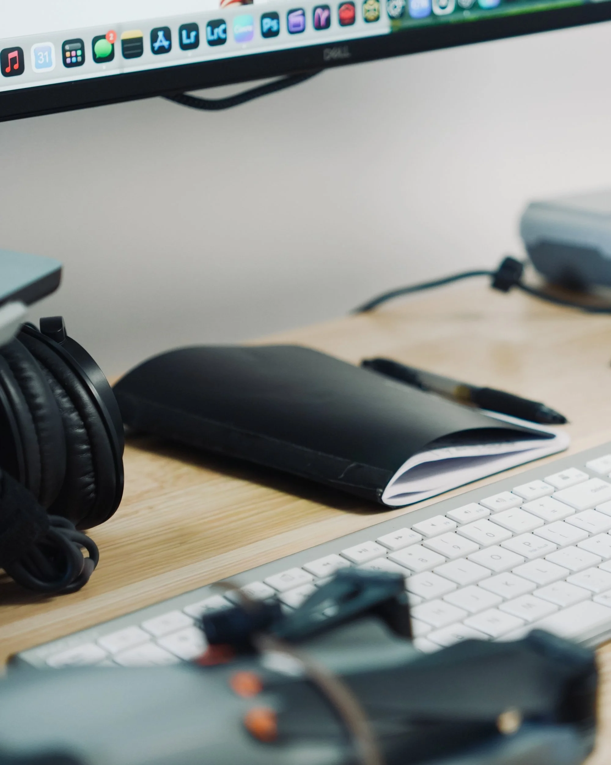 Close-up image of a computer keyboard on a desk, with headphones and a notebook with a pen in the background, and a monitor with app icons at the top.