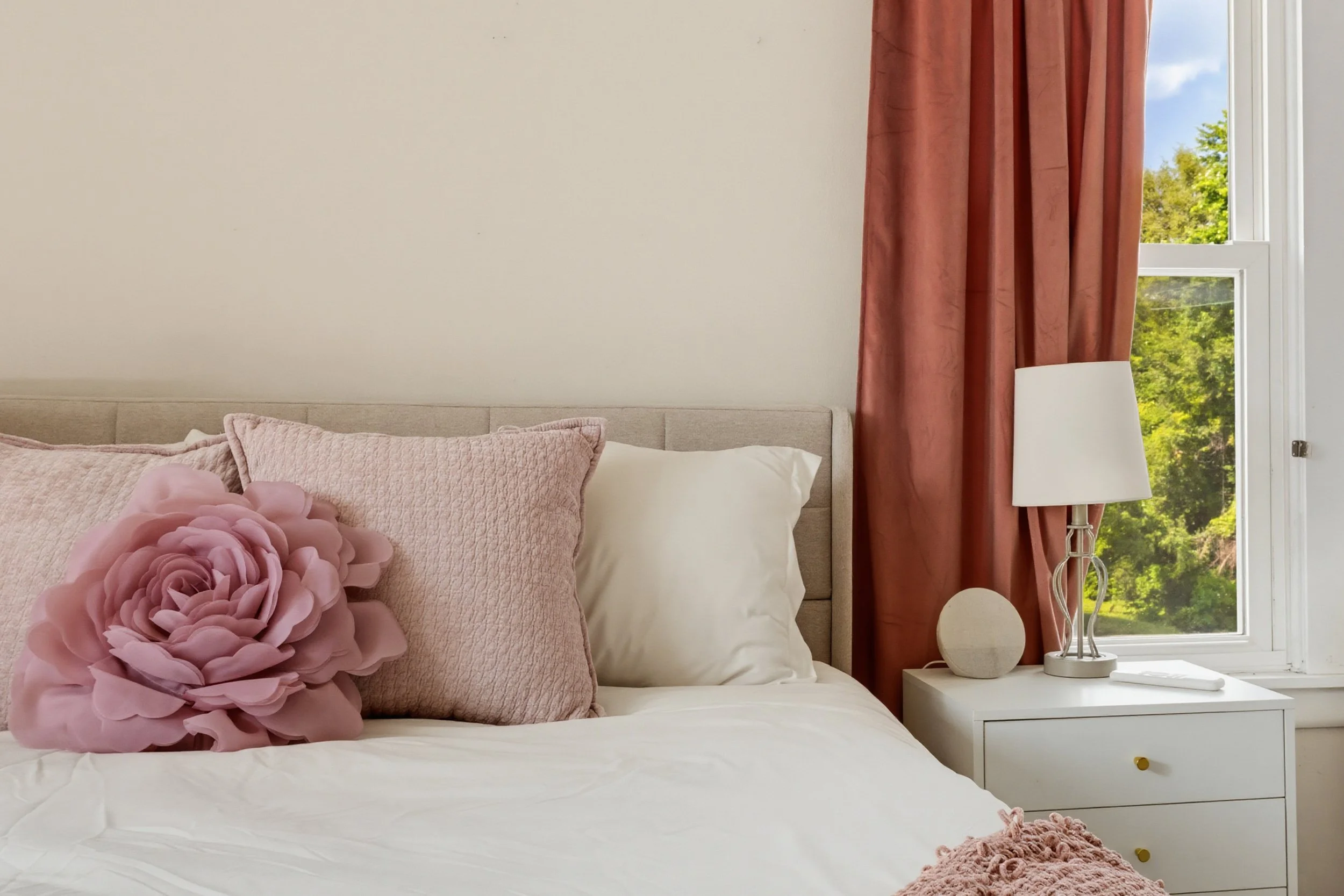 A neatly made bed with pink throw pillows and a decorative pink flower pillow, surrounded by a white bedside table with a white lamp, next to a window with red curtains and green trees outside.