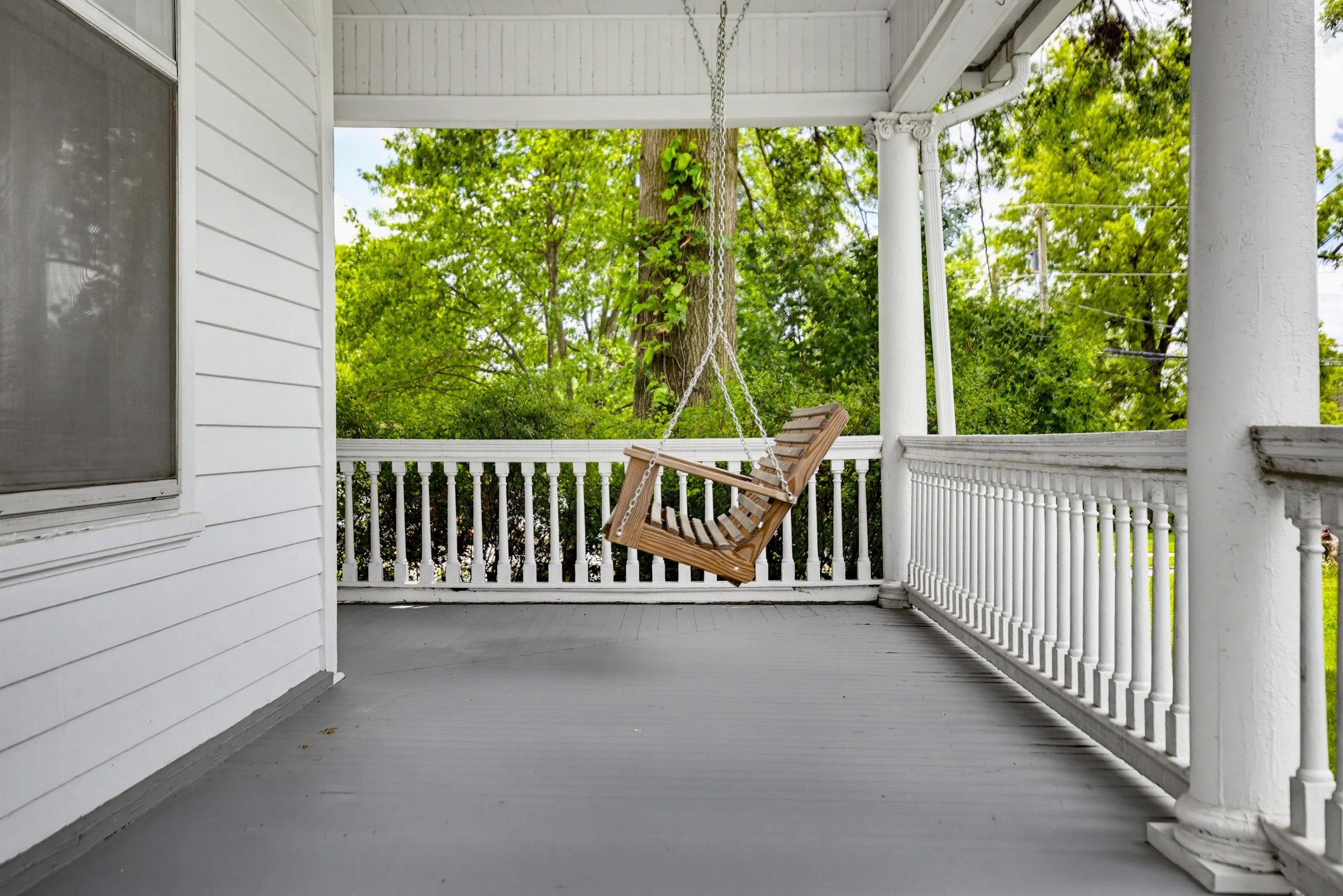 Porch with a wooden swing hanging from chains, white railings and pillars, surrounded by green trees.