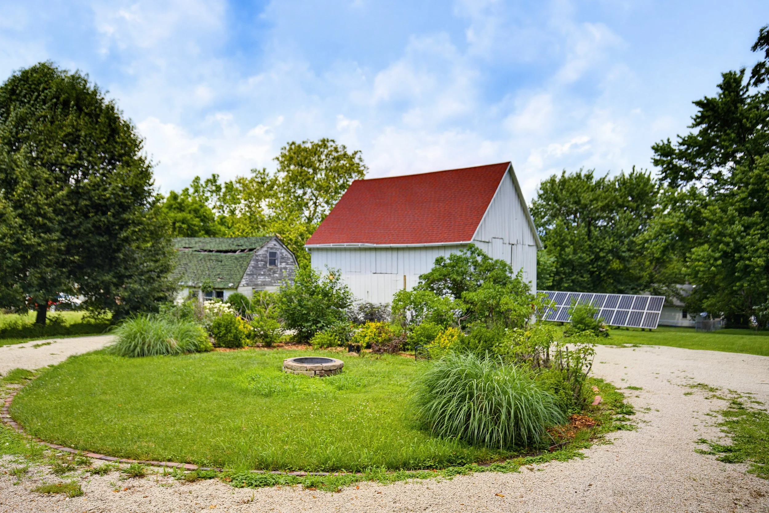 A peaceful backyard garden with a green lawn, trees, and bushes. There is a white building with a red roof in the background and solar panels on the ground. A small circular fire pit is in the center of the garden.
