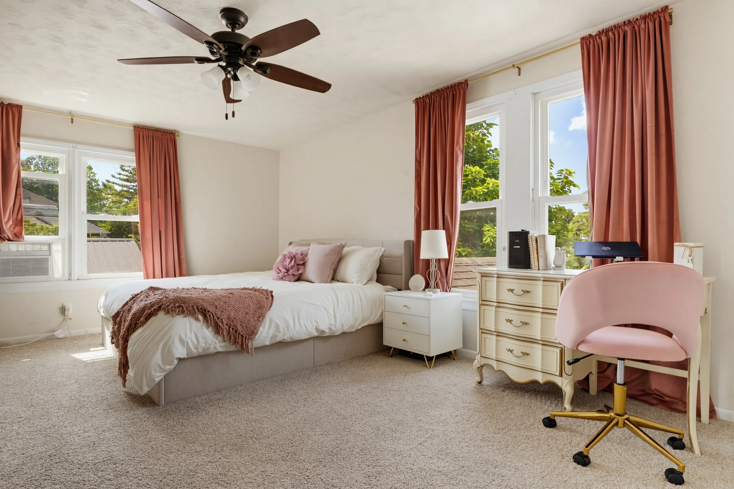 A cozy bedroom with a large window revealing trees outside, a white bed with pillows and a pink throw blanket, pink curtains, a white nightstand with a lamp, a vintage-style dresser, and a pink office chair on a beige carpet.