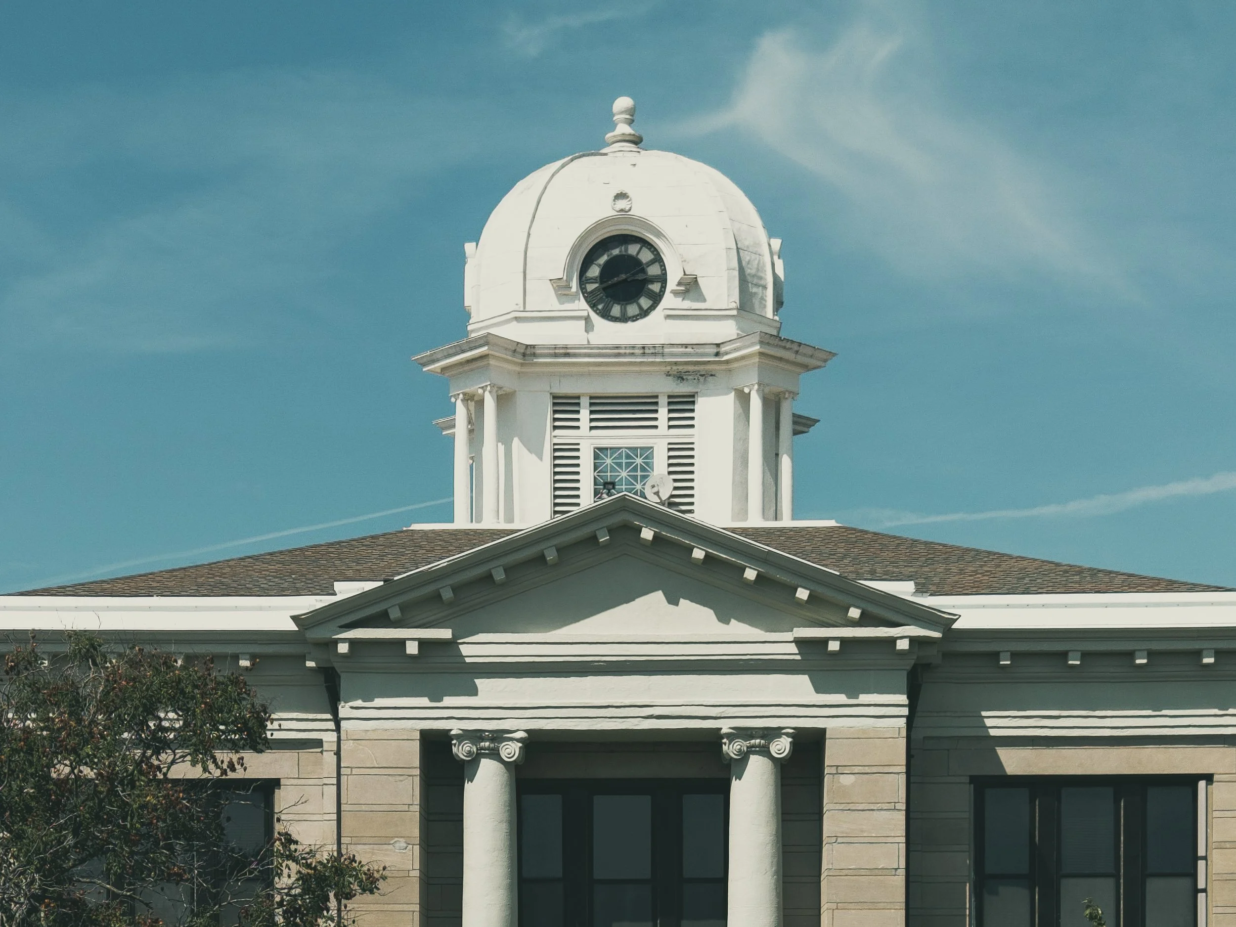 A white clock tower with a domed roof and a clock face showing the time. The tower has architectural details and is part of a larger building with a peaked roof. The sky in the background is mostly clear with some light clouds.