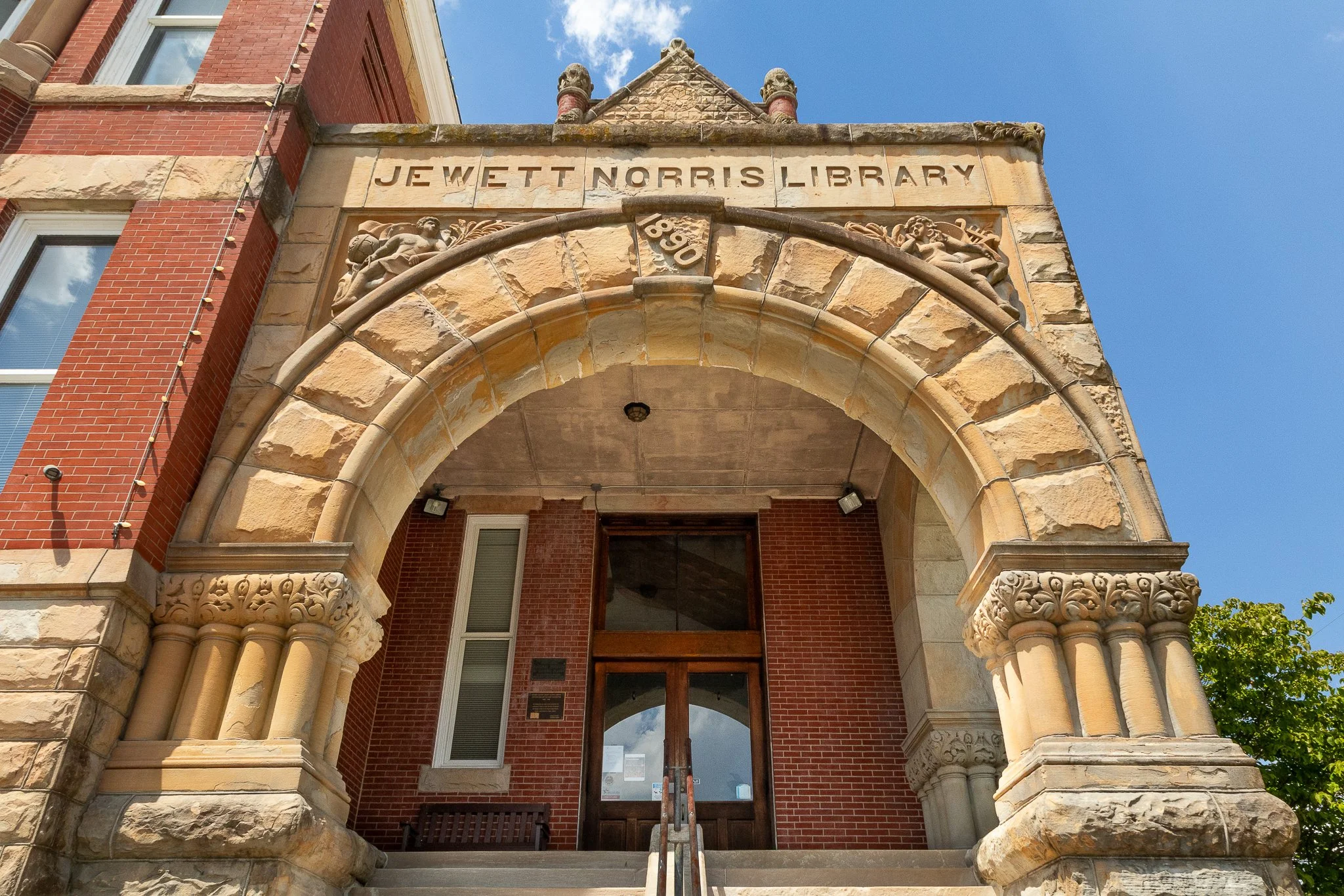 The entrance of the Jewett Norris Library, built with a stone archway and decorative carvings, featuring the library's name on a stone plaque, with a red brick building behind and a blue sky overhead.
