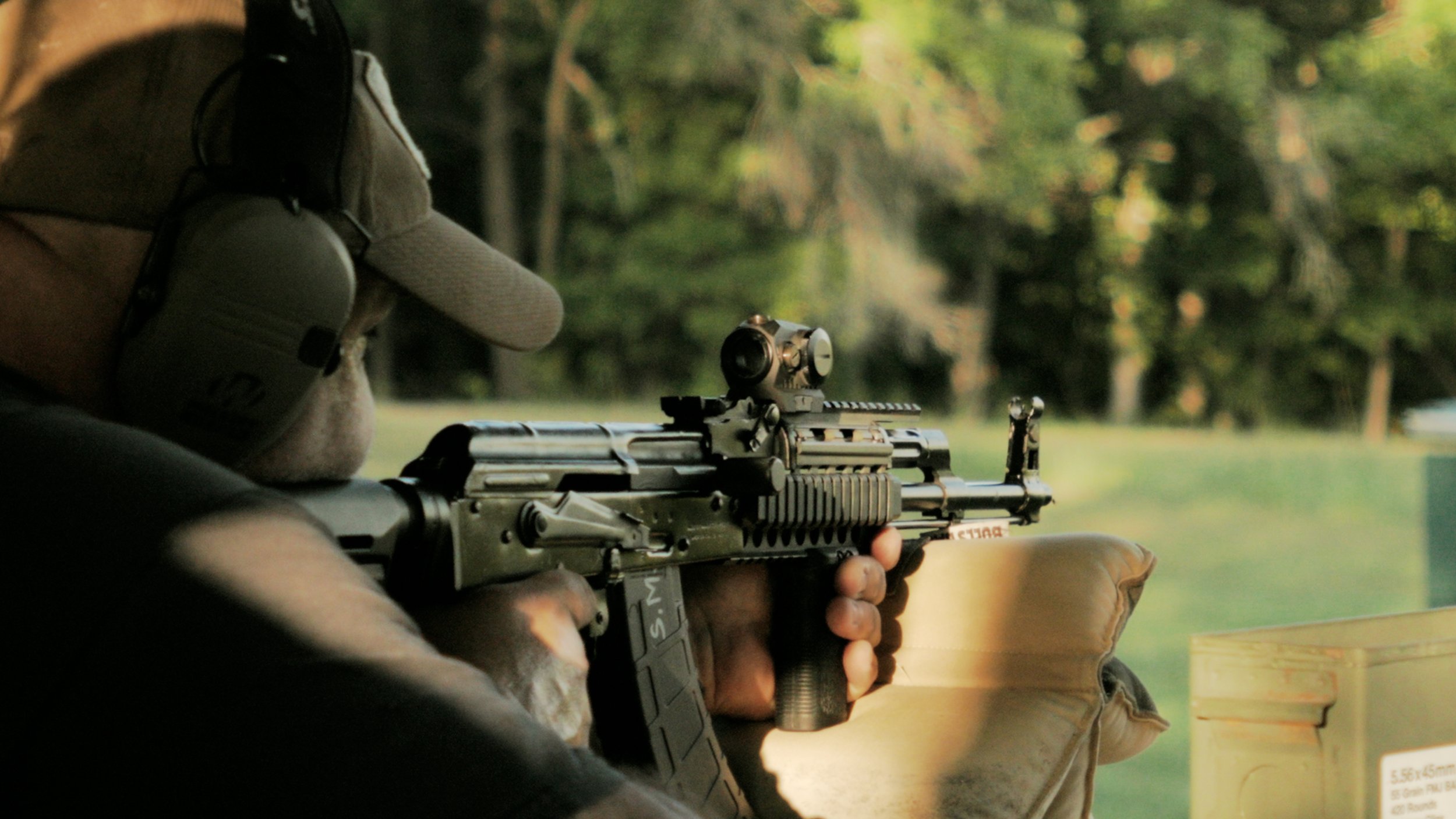 Person aiming a rifle outdoors, wearing camouflage gloves and beige clothing, with a blurred background of trees and greenery.