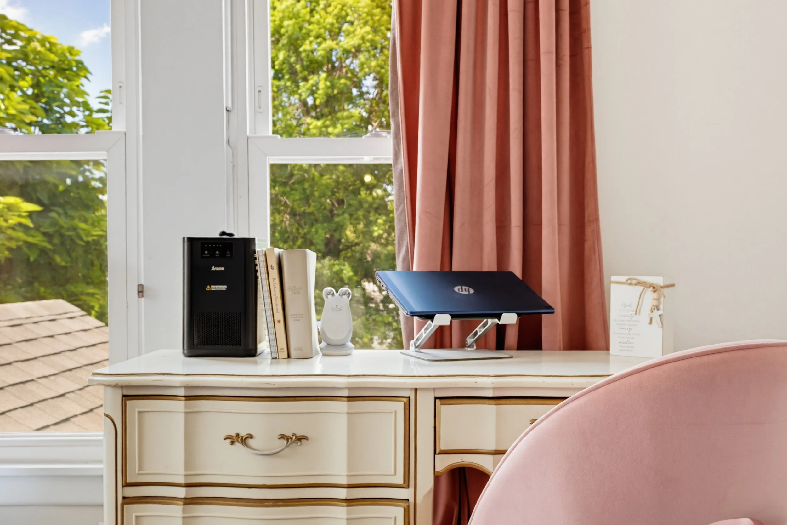 A white desk with gold accents in front of a window with pink curtains. On the desk are a black printer, a stack of books, a white ceramic animal figure, a laptop, and a framed note card. Outside the window, green trees and a rooftop are visible.