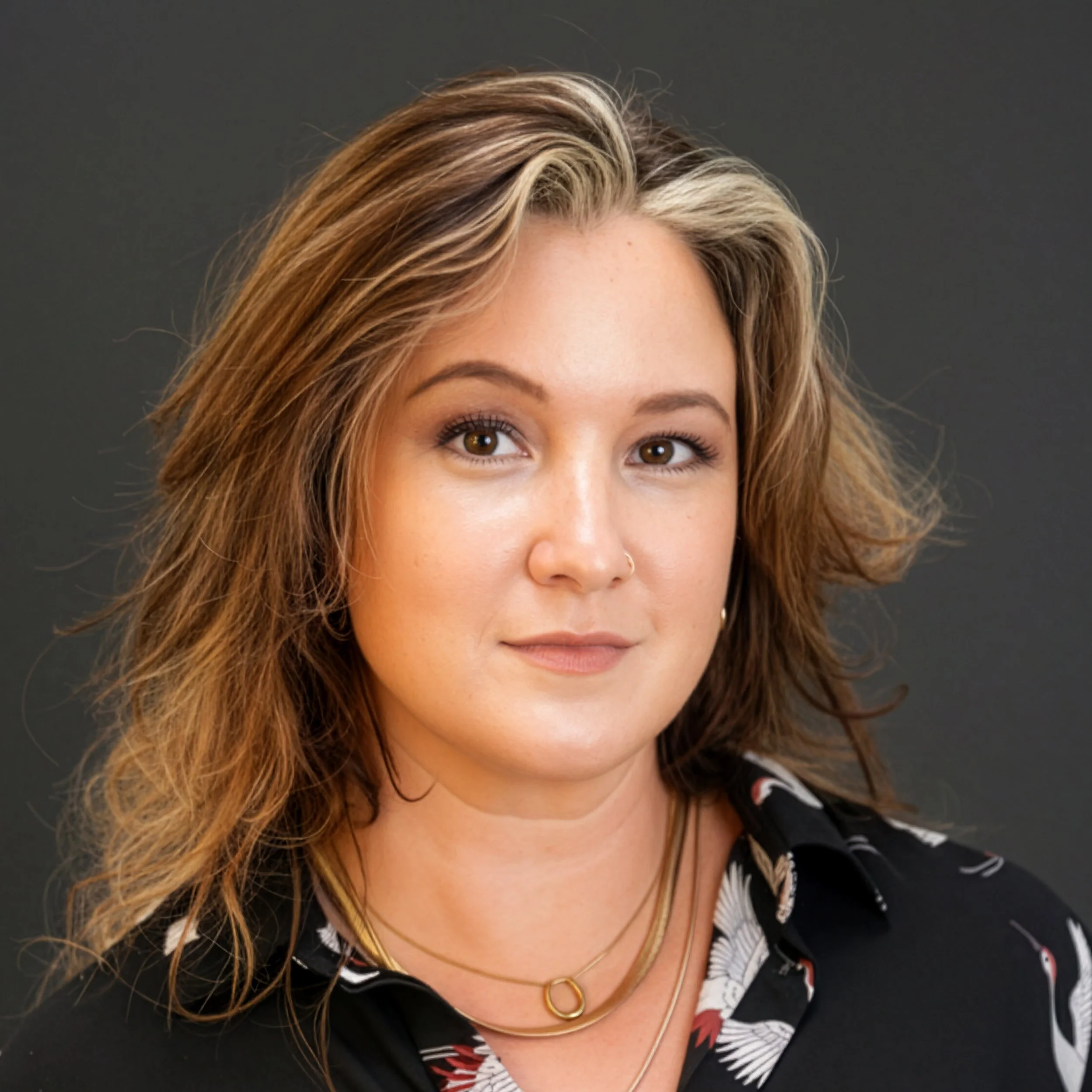 A woman with short brown hair, tattoos on her arms, wearing a tiger print shirt and ripped jeans, sitting on a green chair with her hands on her cheeks, smiling at the camera against a white background.