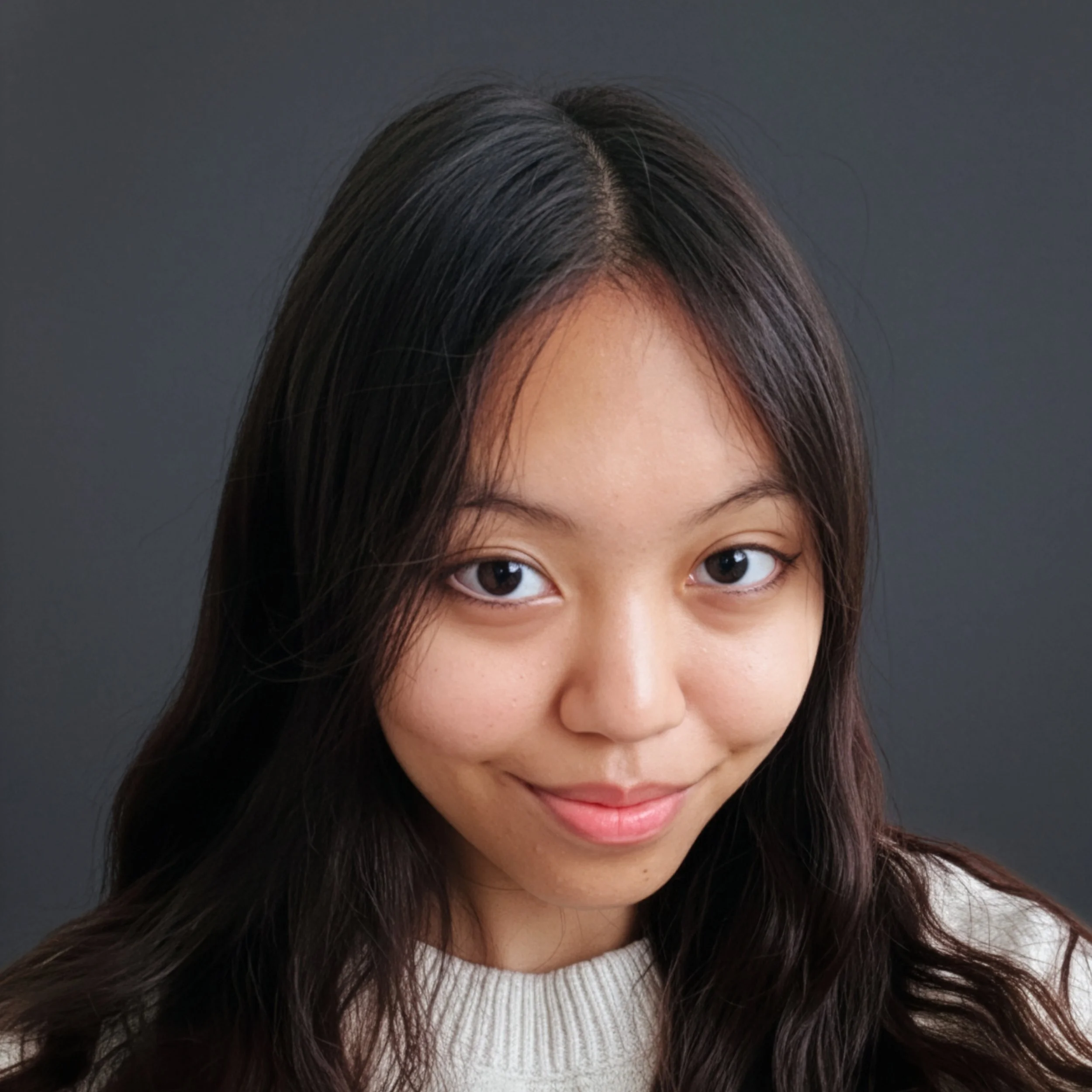 Close-up portrait of a woman with long dark hair, smiling slightly, wearing a white sweater, against a dark gray background.