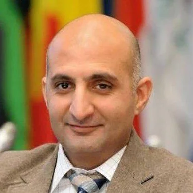 A bald man in a brown suit and tie smiling, with a colorful blurred background.