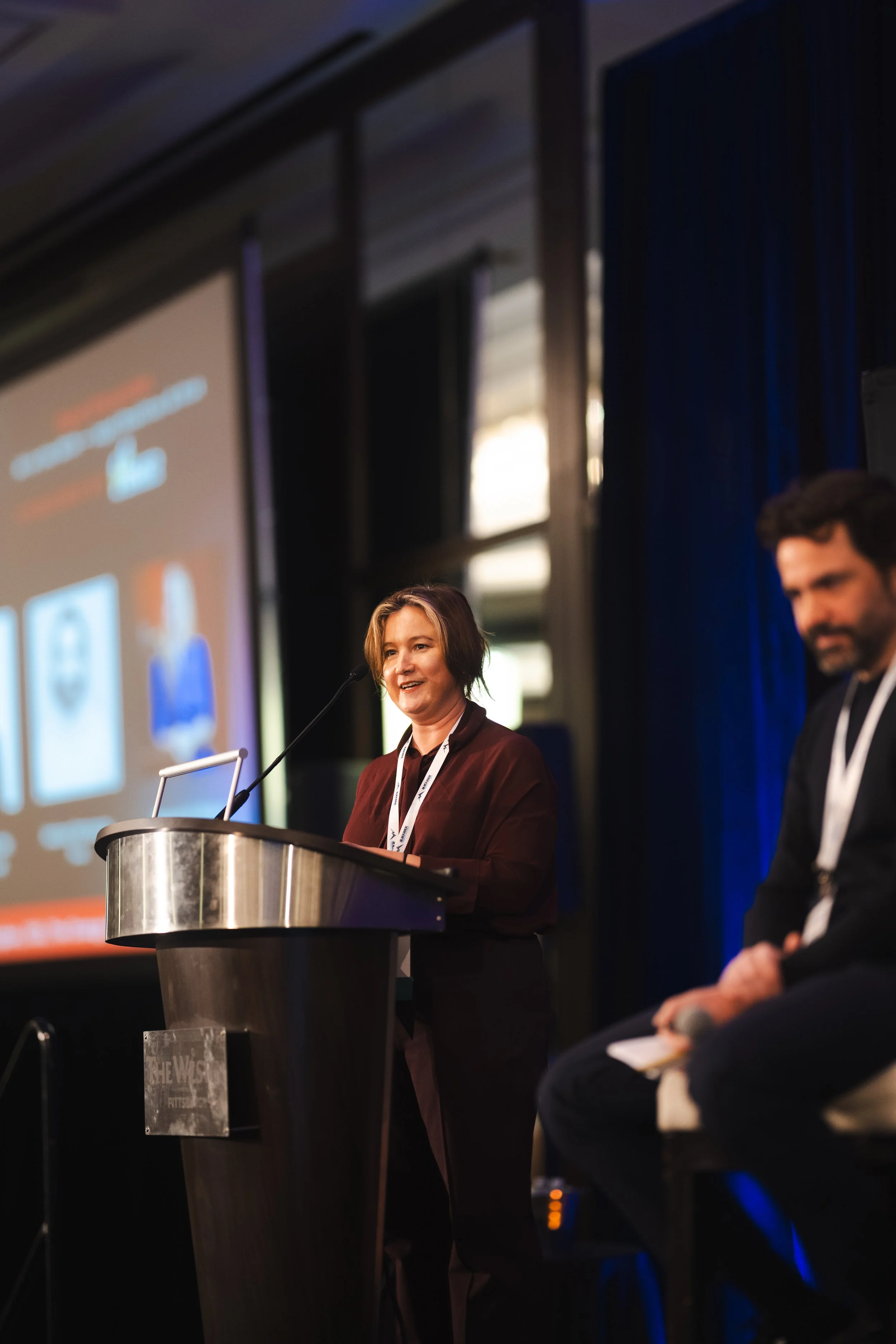 A woman speaking at a podium during a conference or event with a presentation slide projected in the background, and a man sitting nearby.