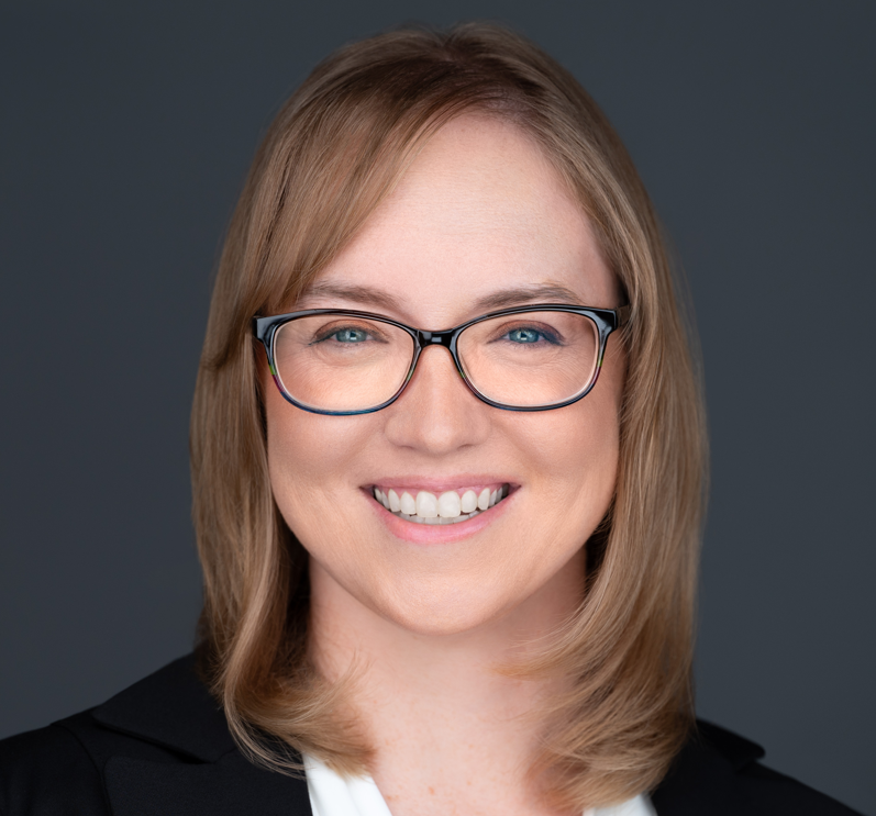 A woman with light brown hair and glasses smiling in front of a dark background.