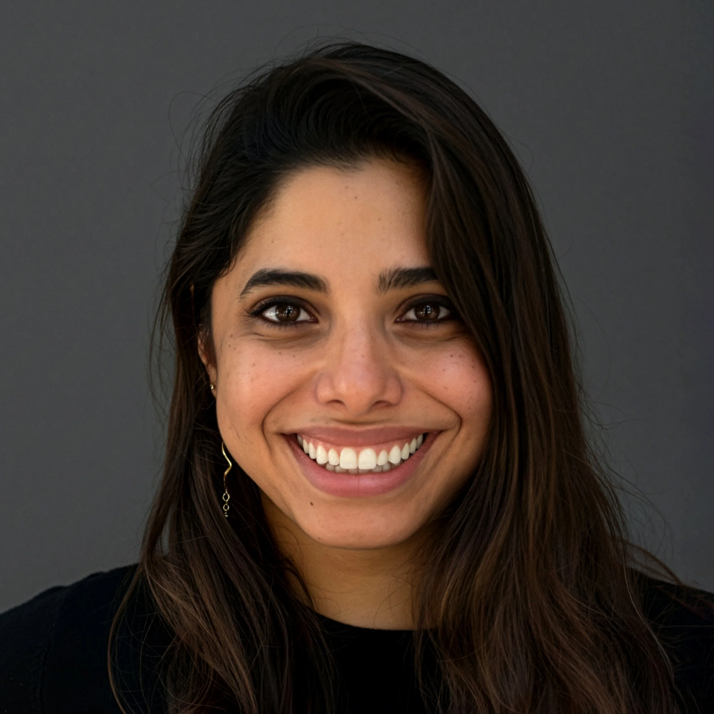 A woman with long, wavy dark hair and a bright smile, wearing a black top, posing outdoors with a rustic wooden background.