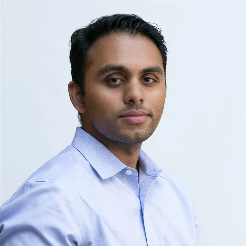 A confident man with short black hair wearing a light blue button-up shirt, posing against a plain light background.