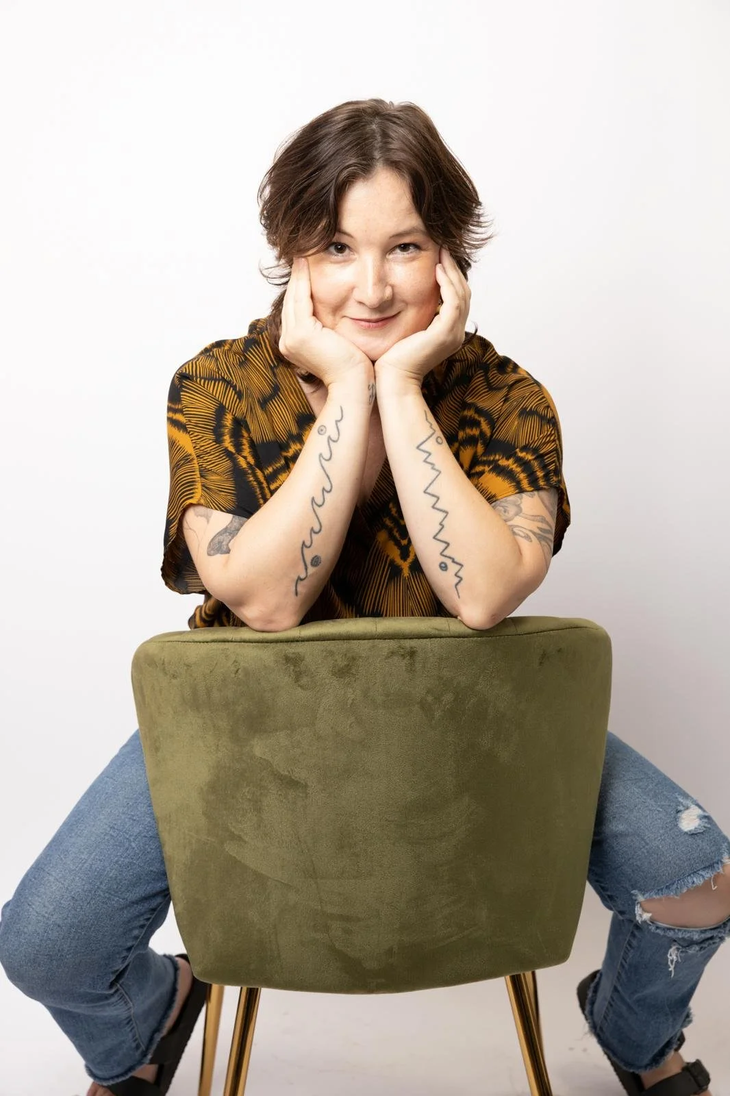 A woman with short brown hair, tattoos on her arms, and a playful expression, sitting on a chair with a green backrest, leaning forward with her chin resting on her hands.