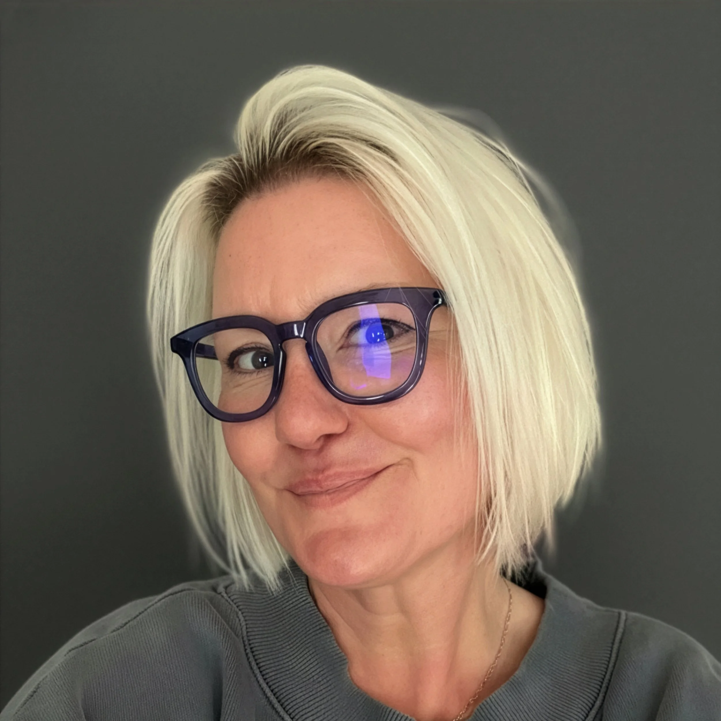 Partial portrait of a woman with blonde hair wearing glasses and a gray top, against a dark background.