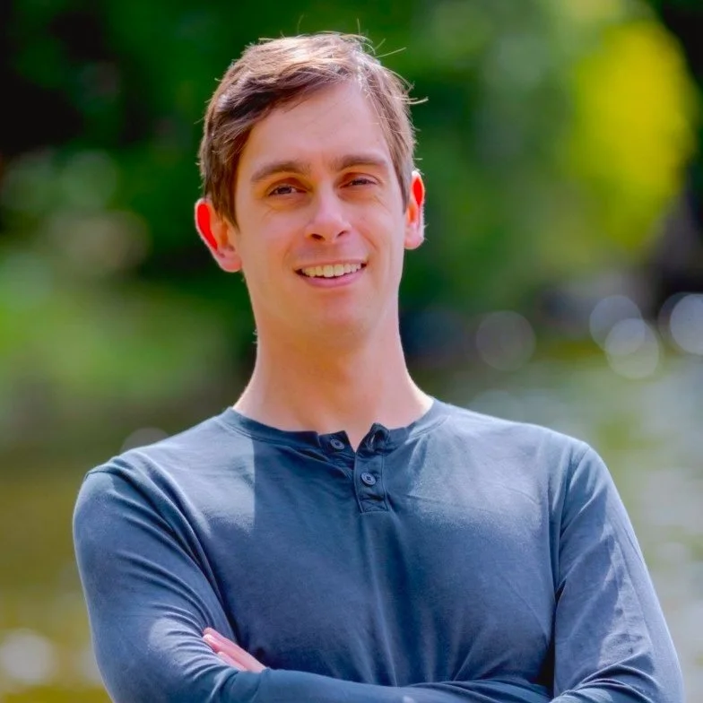 A young man smiling outdoors with arms crossed, wearing a long-sleeve dark blue shirt, with blurred greenery and water in the background.