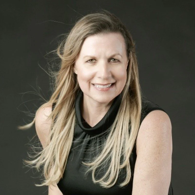 A woman with long blonde hair, smiling, wearing a black sleeveless top, against a dark background.