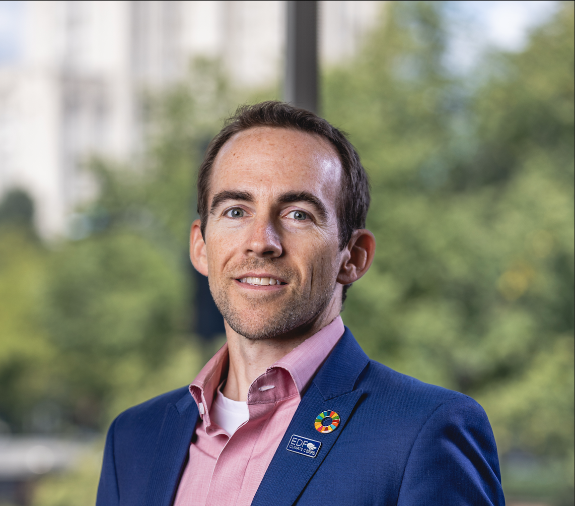 Professional man in pink shirt and dark blazer with a multicolored round pin and an EDF badge, standing indoors with a window and blurred green outdoor trees in the background.