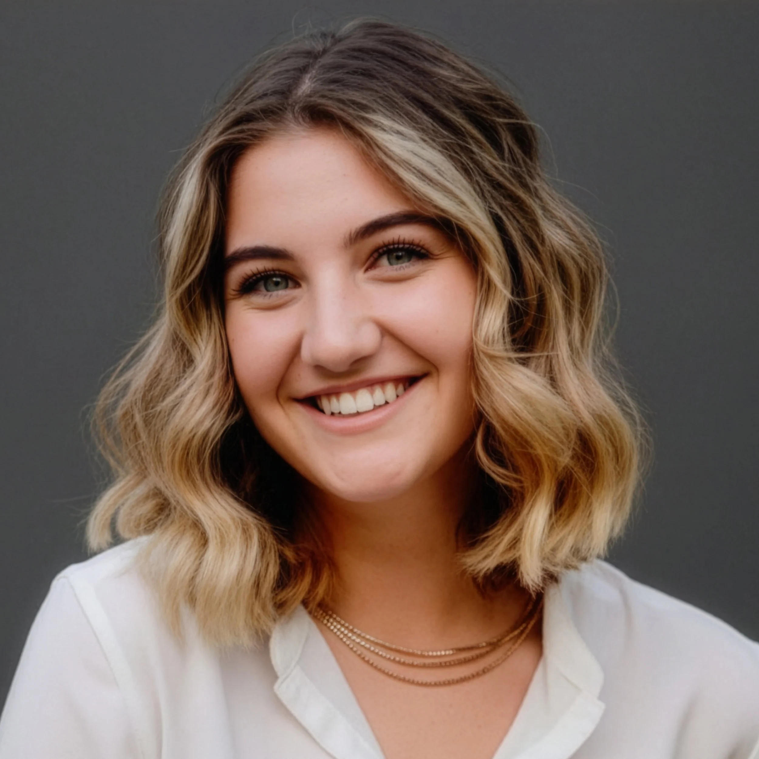 Close-up of a smiling woman with shoulder-length, wavy blonde hair wearing a white top and layered necklaces against a gray background.