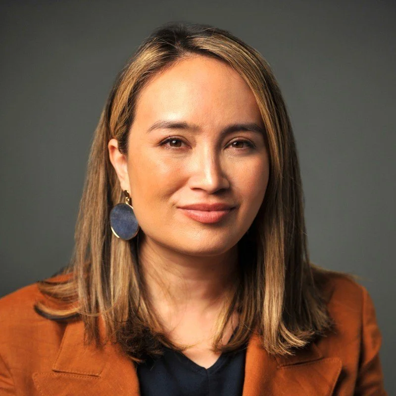 A woman with shoulder-length brown hair, wearing a brown blazer over a navy top, and large dark earrings, smiling softly at the camera against a gray background.