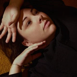 Een close-up van een jonge vrouw met een lichte huid, donker haar en een zwart shirt, die op haar zij ligt met haar hoofd op haar hand.