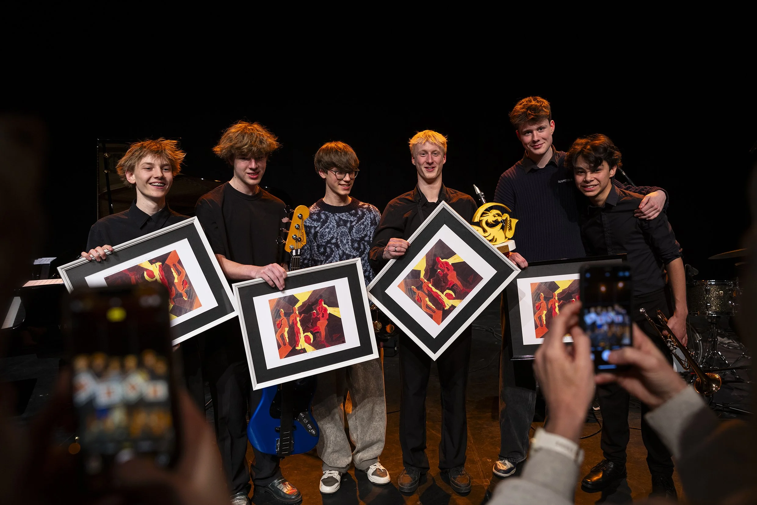 Een groep jonge mannen staat op een podium met kunstwerken en een trofee, terwijl mensen in de zaal foto's maken.