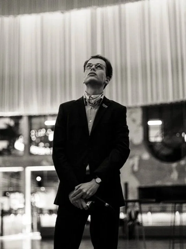 Een man in een zwart pak met een stropdas en een horloge, met een glas in zijn hand, kijkt omhoog in een grote hal of zaal.