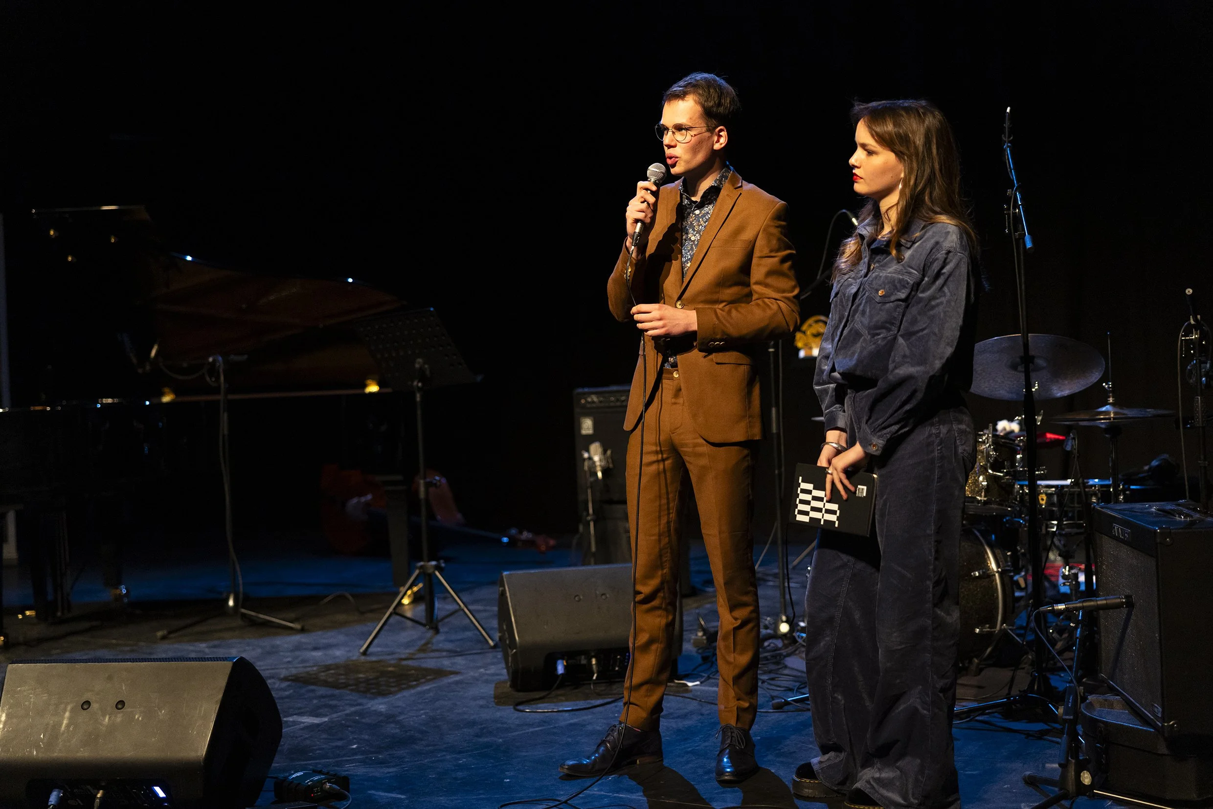 Twee mensen staan op een podium met muziekinstrumenten op de achtergrond. Eén persoon houdt een microfoon vast en spreekt, de andere kijkt toe.