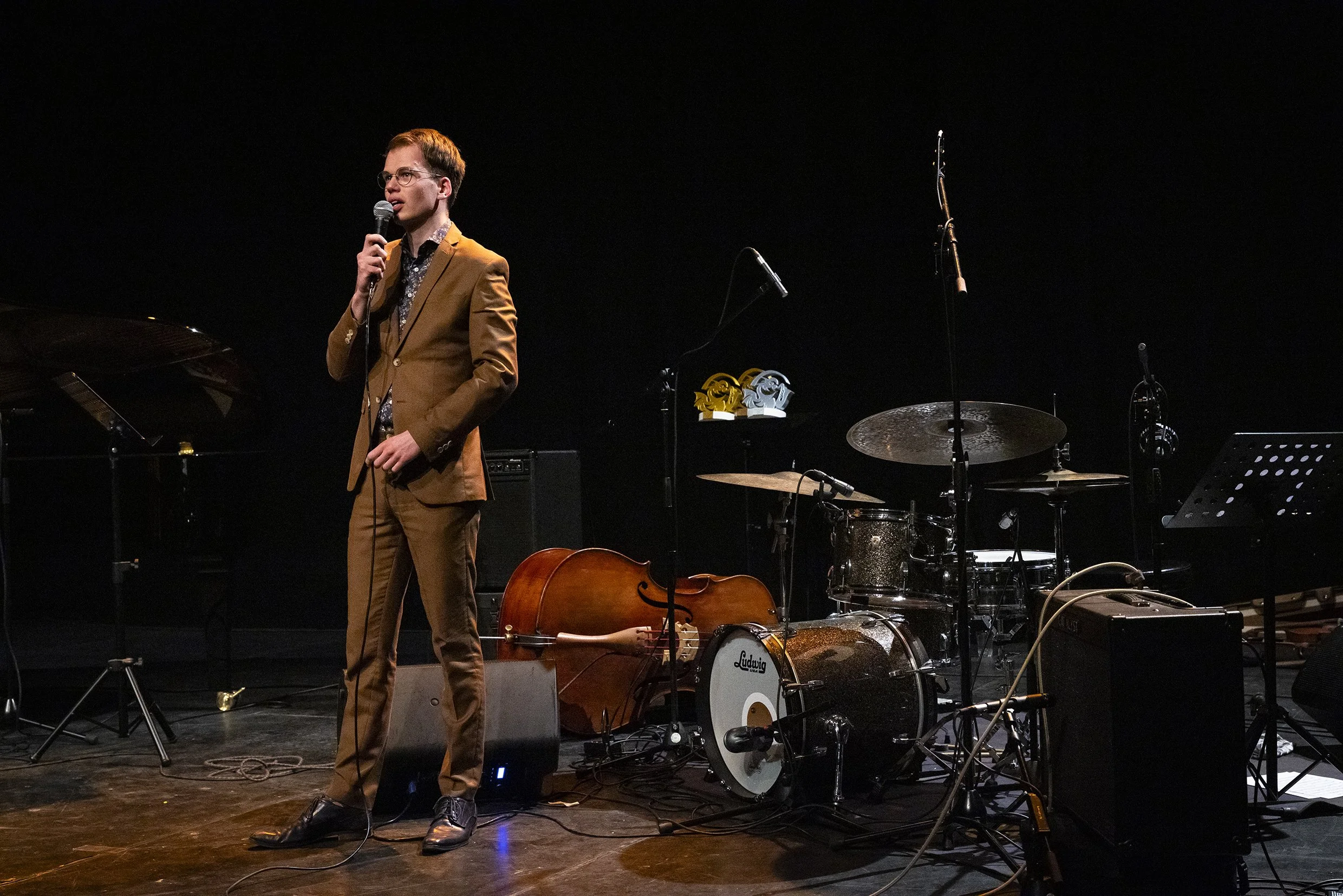 Een man in een bruin pak staat op een podium en houdt een microfoon vast, omringd door muziekinstrumenten zoals een drumstel en een cello.