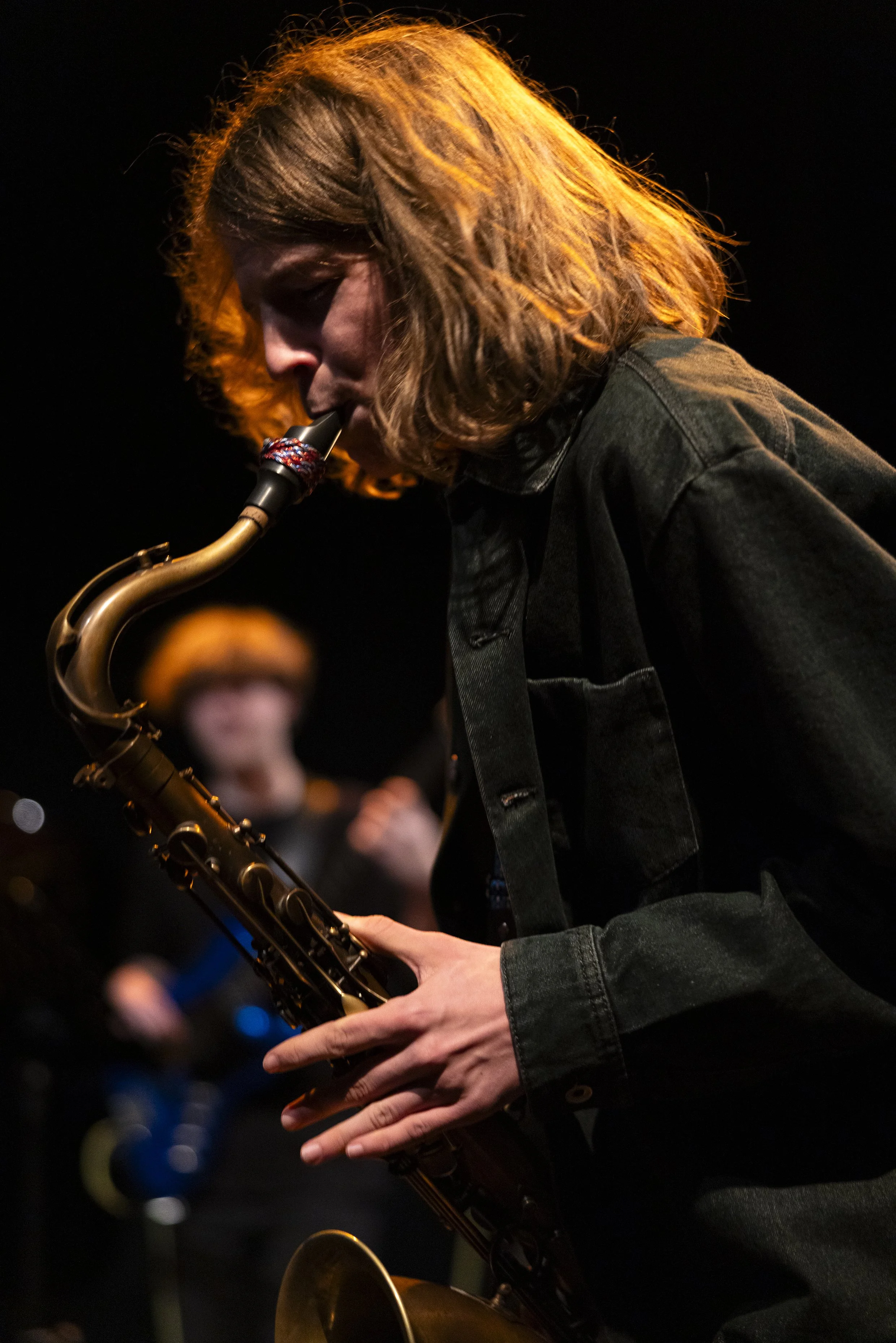 Een vrouwelijke saxofonist speelt op een podium, met donkere achtergrond en warme lampverlichting op haar haar en instrument.