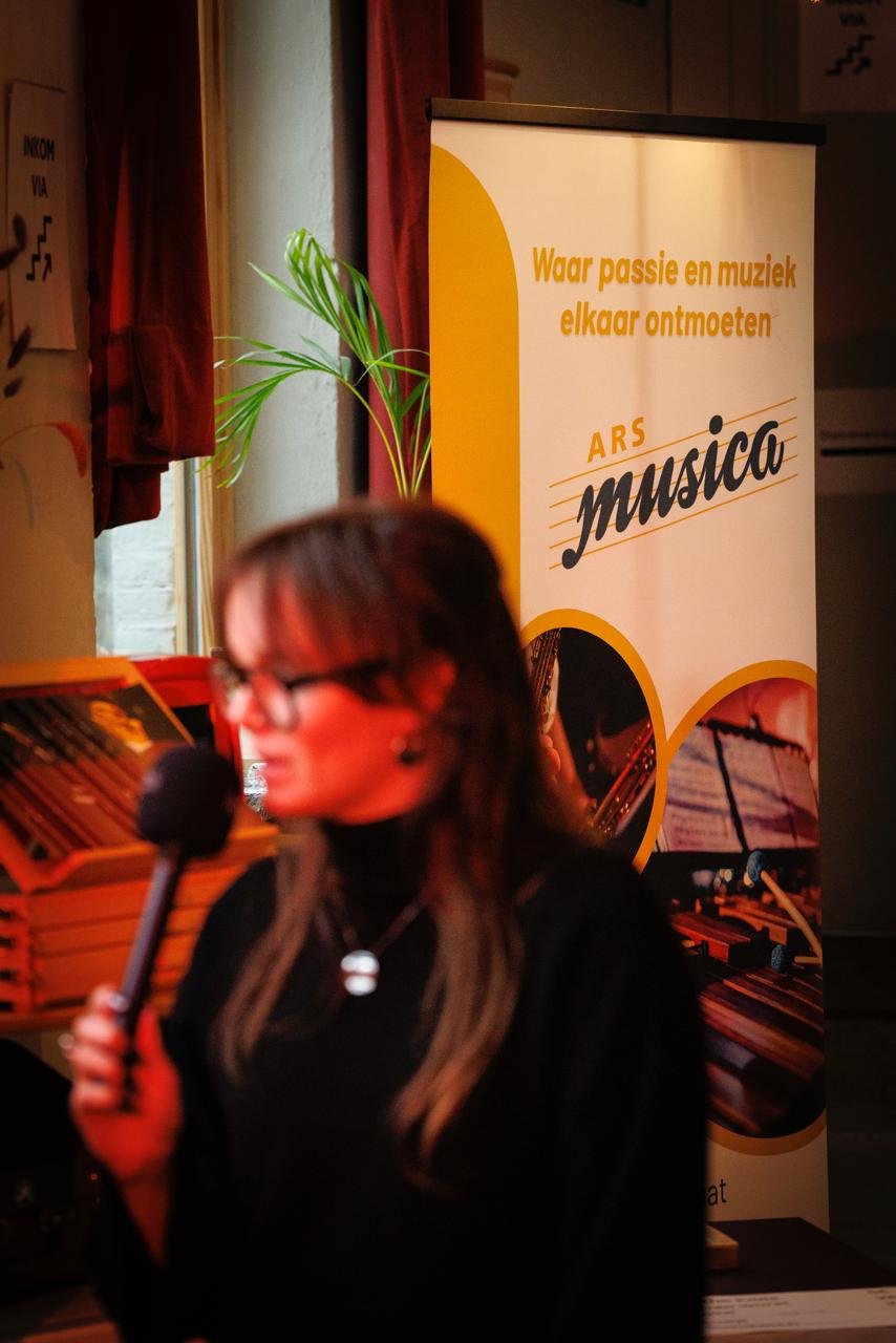 Een vrouw met bril houdt een microfoon vast, op de achtergrond een verticale banner met tekst over muziek en een afbeelding van een piano.