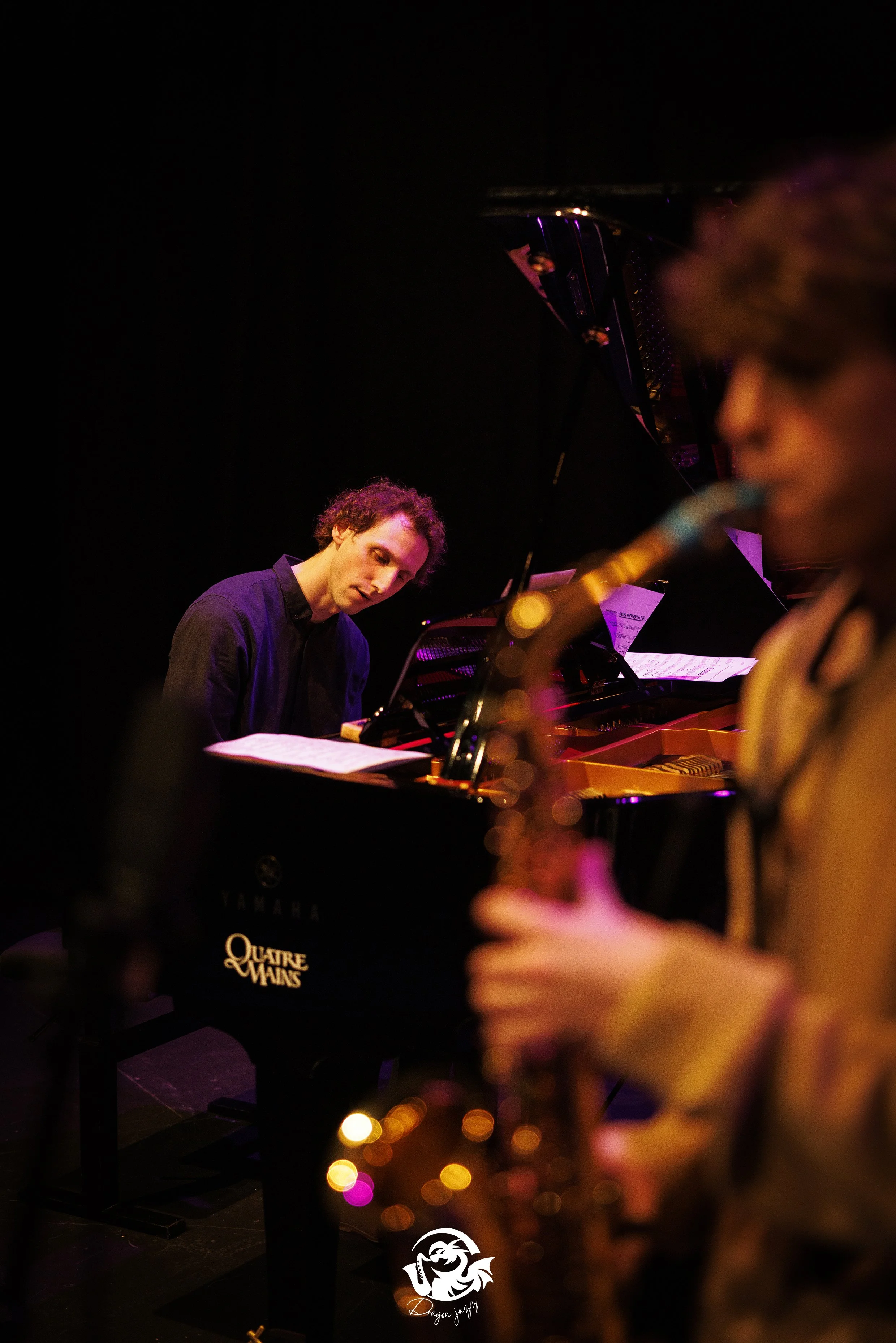 Een jonge man speelt piano op een donkere podium, met een warme, artiestenachtige sfeer. Op de voorgrond is een gezamenlijke jazz-instrument, mogelijk een saxofoon, vaag zichtbaar. Vooral de man en de piano zijn duidelijk zichtbaar, terwijl de rest vaag en kleurrijk verlicht is.