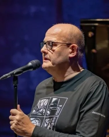 Man met bril en een zwarte T-shirt met grafisch ontwerp, staat voor een microfoon, op een podium met een blauwe achtergrond.