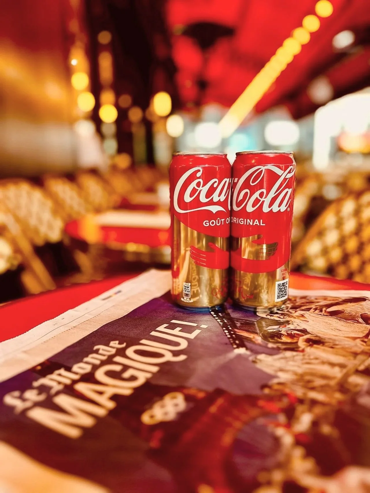 Two cans of Coca-Cola on a table in a brightly lit restaurant or cafe with red and yellow lighting, with blurred background of chairs and tables. Campaign created by Katy Stanage