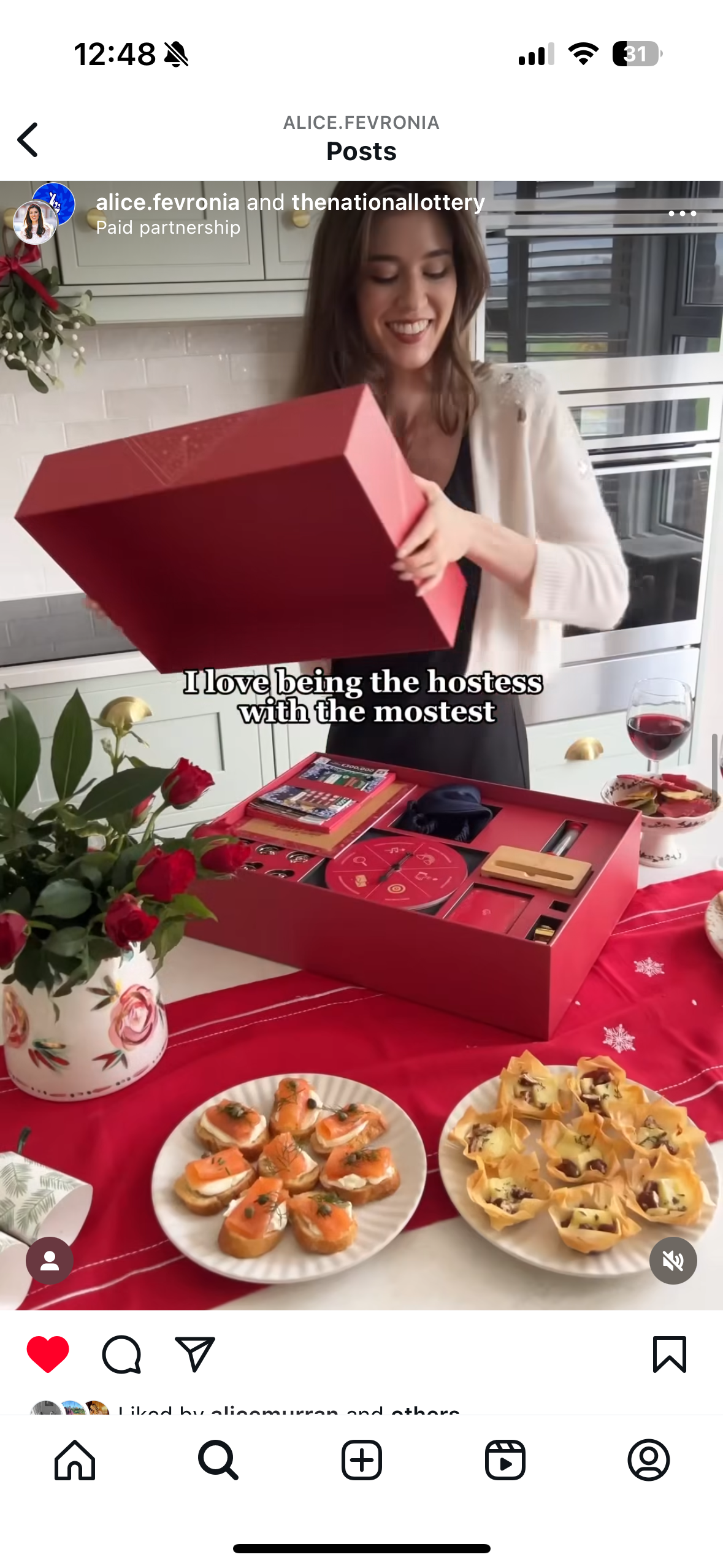 A woman opening a red gift box on a festive table with appetizers, a glass of red wine, and a bouquet of red roses in a patterned vase.