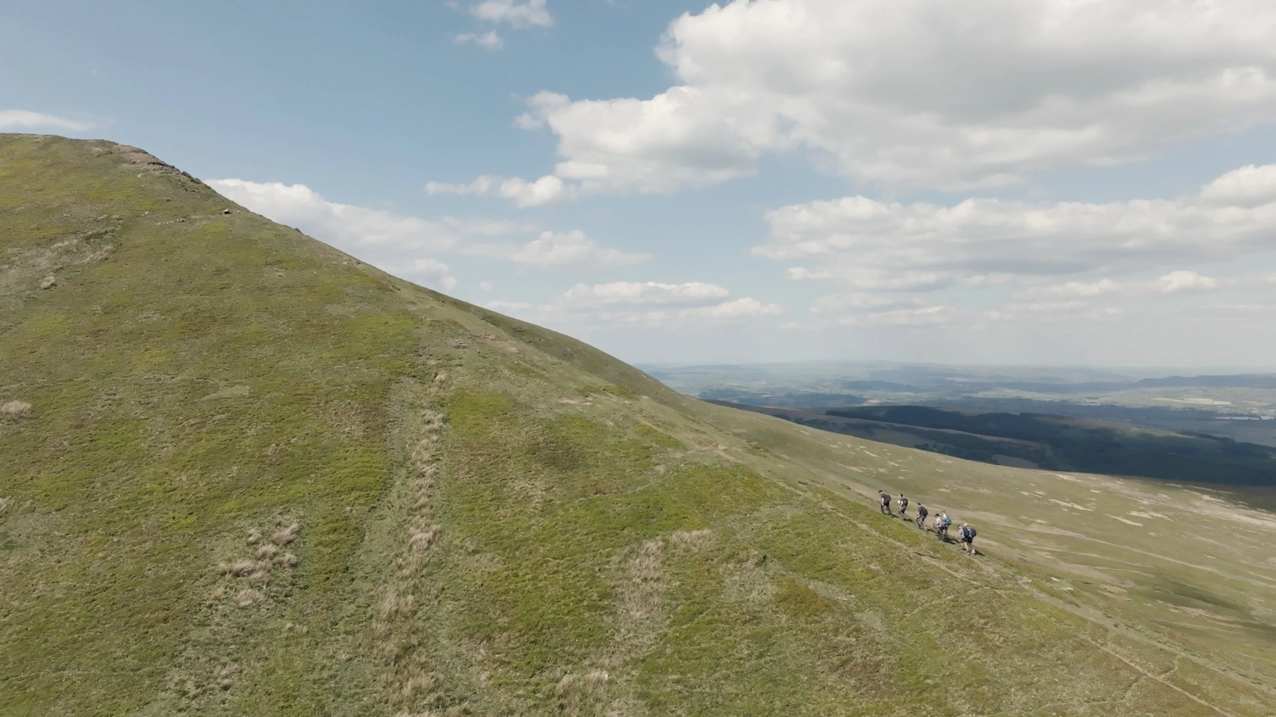 Hiking trail on a green hillside with a group of hikers walking in single file towards the right, under a partly cloudy sky.