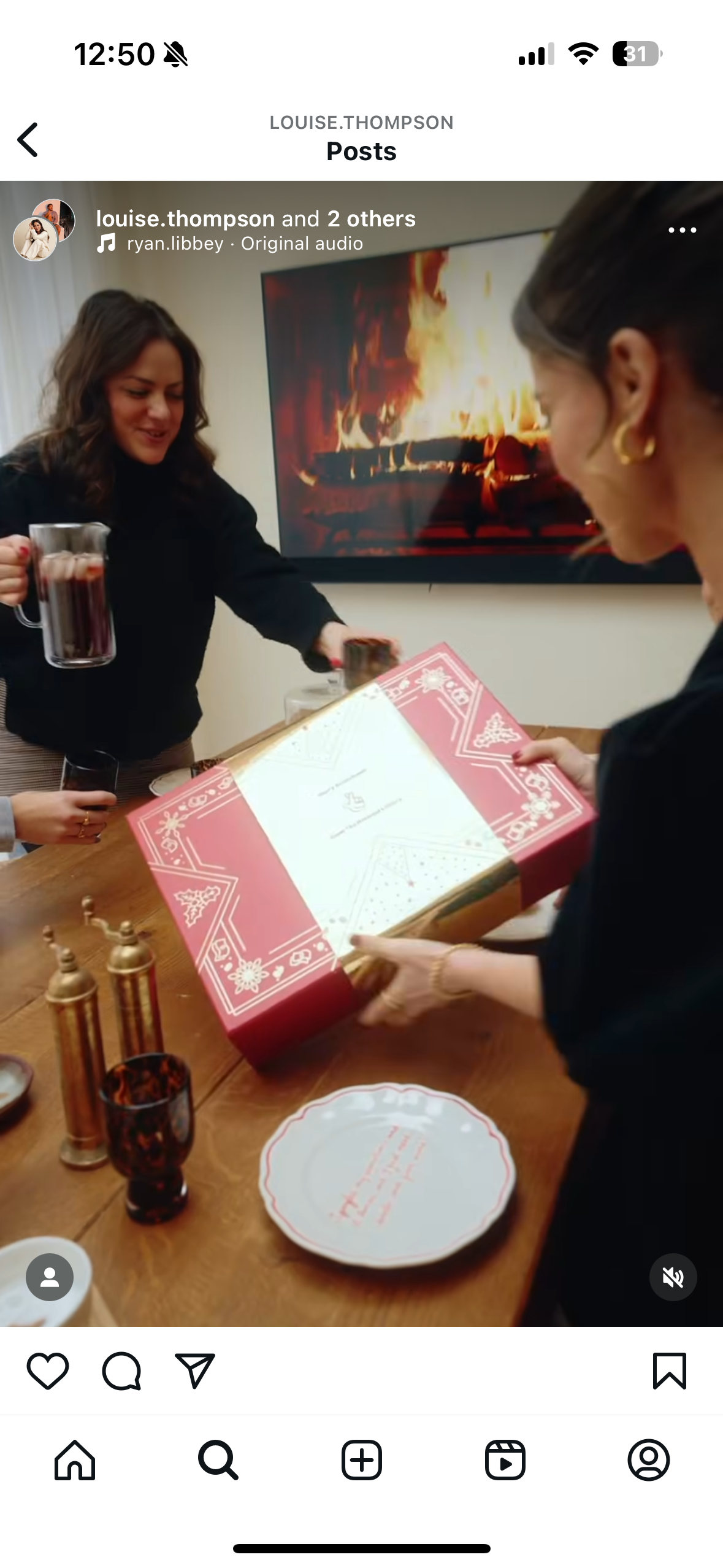 A group of women are gathered around a wooden table with holiday decorations, including a red and white box, glassware, and salt and pepper shakers. One woman is holding a mug of dark beverage, and another is opening a gift box decorated with holiday patterns. A flat-screen TV showing a fire in a fireplace is on the wall behind them.