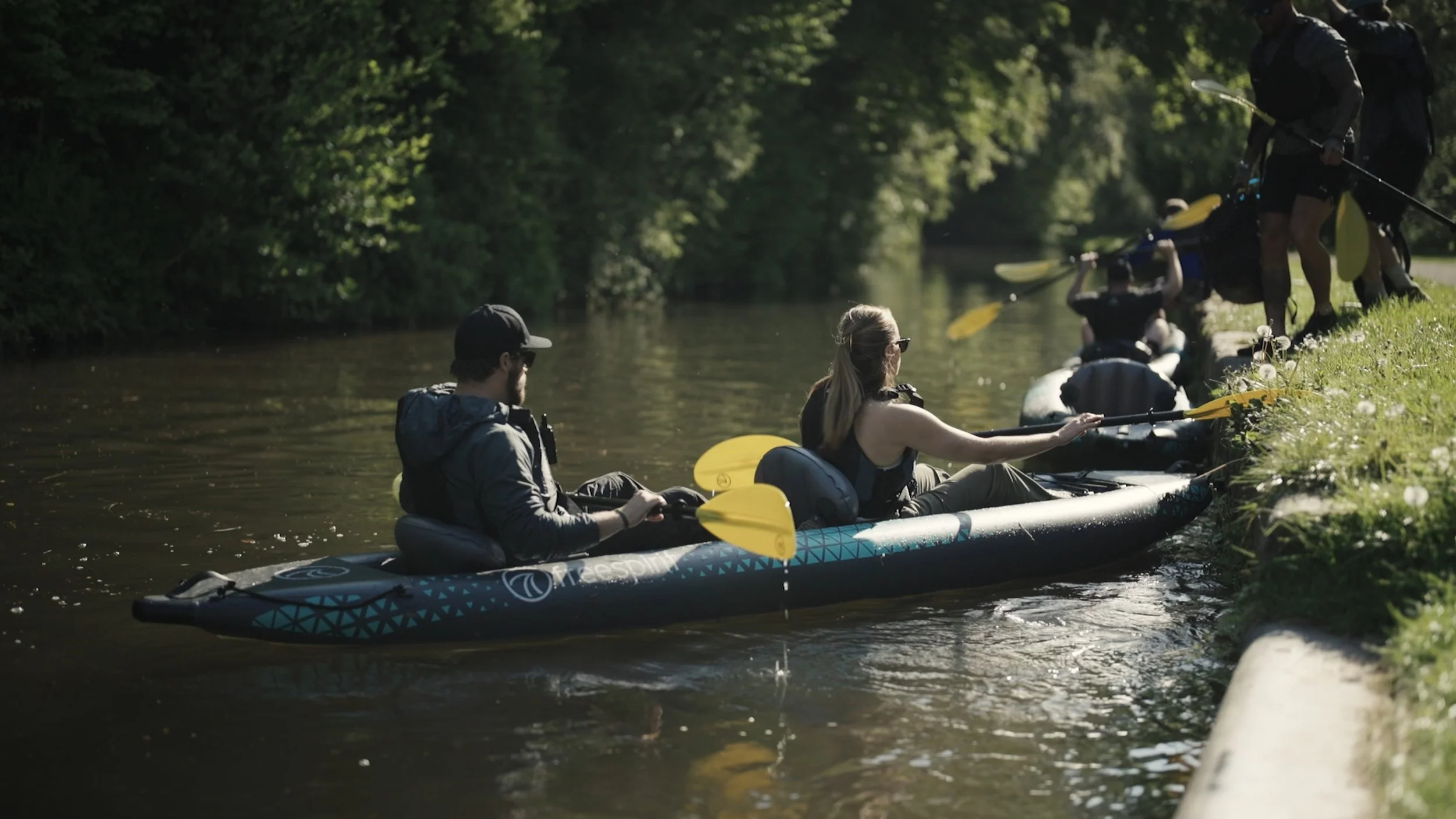 A group of people kayaking on a calm river surrounded by greenery, with some sitting in the kayak and another standing on the riverbank preparing to enter the water.
