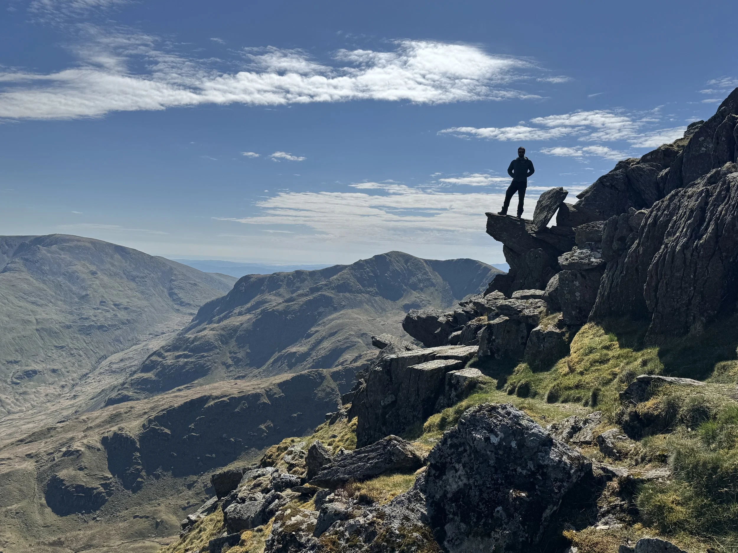 A person standing on a rocky cliff looking out over mountain valleys and peaks under a blue sky with scattered clouds.