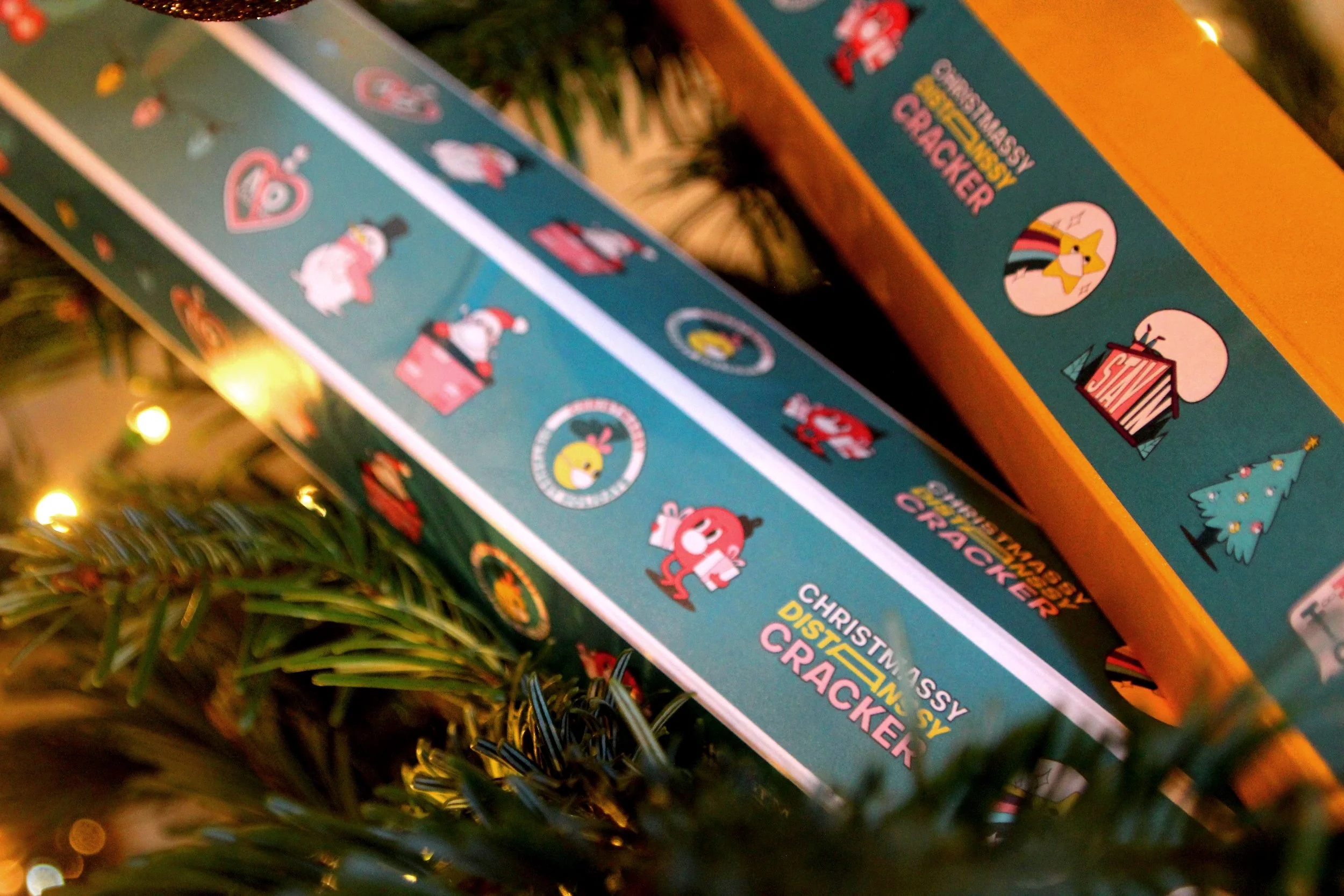 Close-up of a colorful box of Christmas-themed snacks or crackers, decorated with festive stickers including Santa, a pig, a dog, and a Christmas tree, partially obscured by pine branches and yellow string lights. Campaign created by Katy Stanage