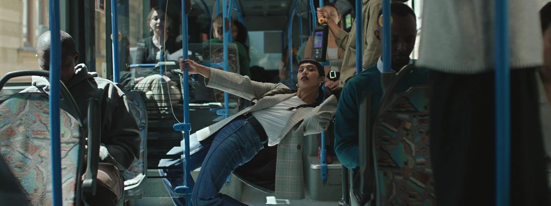 A woman with braided hair is lying sideways on a bus seat, holding a phone, surrounded by other passengers. Campaign created by Katy Stanage