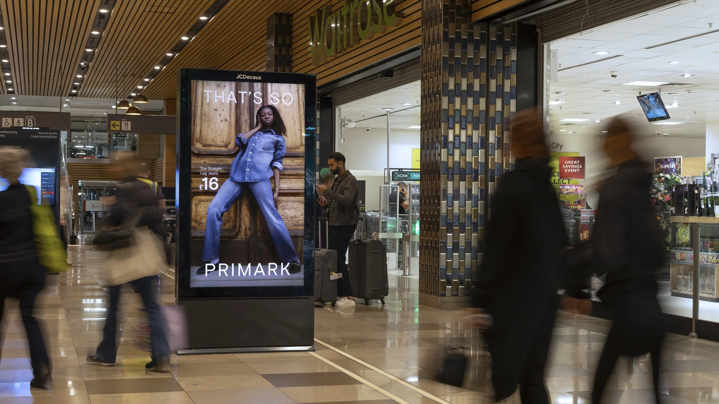 Digital advertisement for Primark featuring a woman in blue jeans and a denim jacket, with text indicating a price of £16, inside an airport terminal.