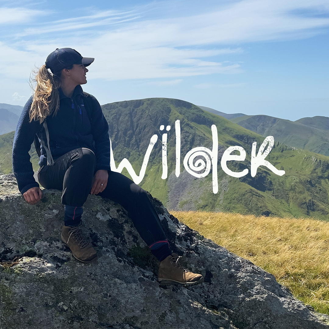 A woman sitting on a rock overlooking green mountains, wearing outdoor hiking gear and a cap, with the word 'wilder' written across the image. Founded by Katy Stanage