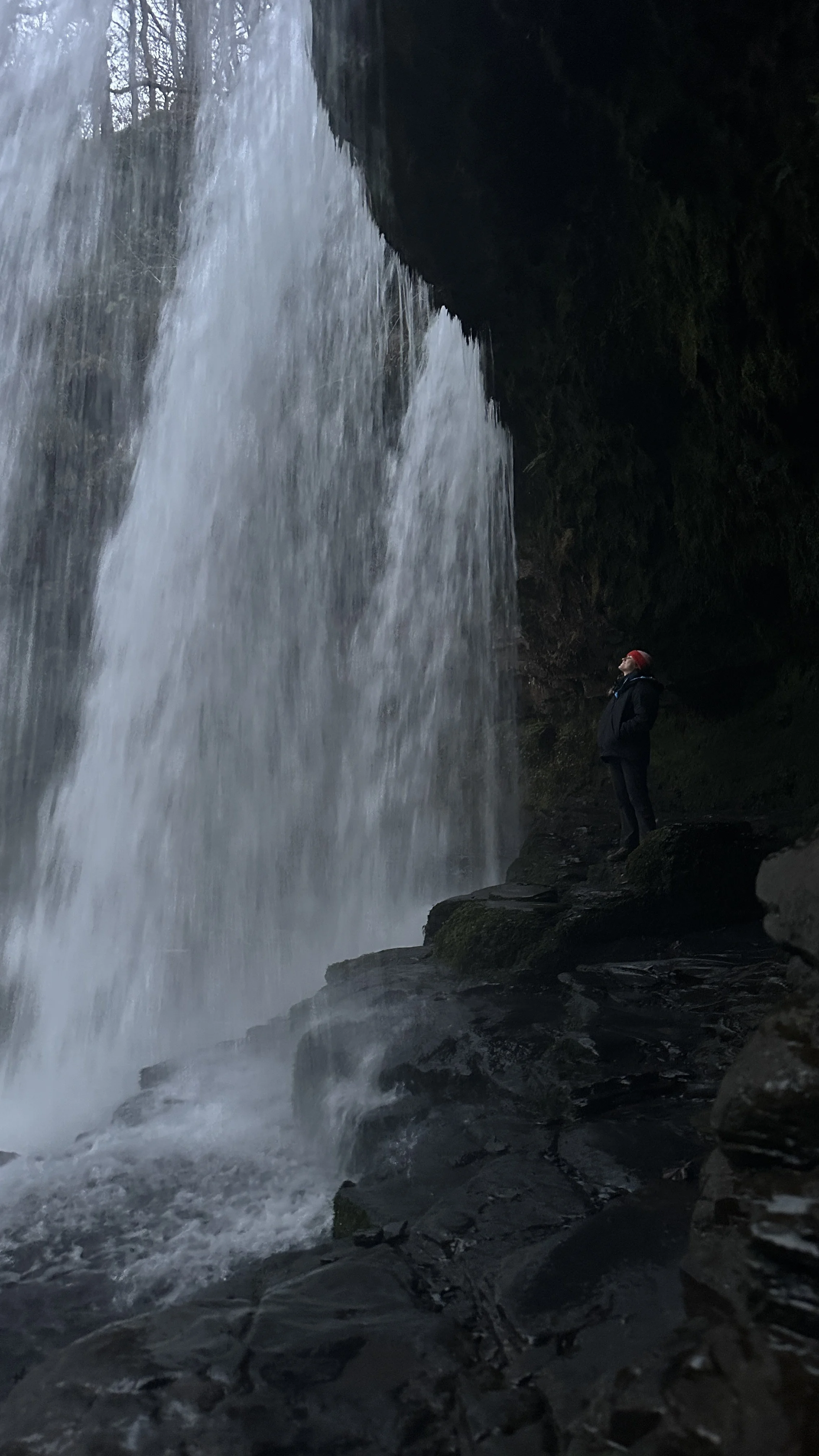 Person in black jacket and red hat standing on rocks near a waterfall, looking up at the falling water.
