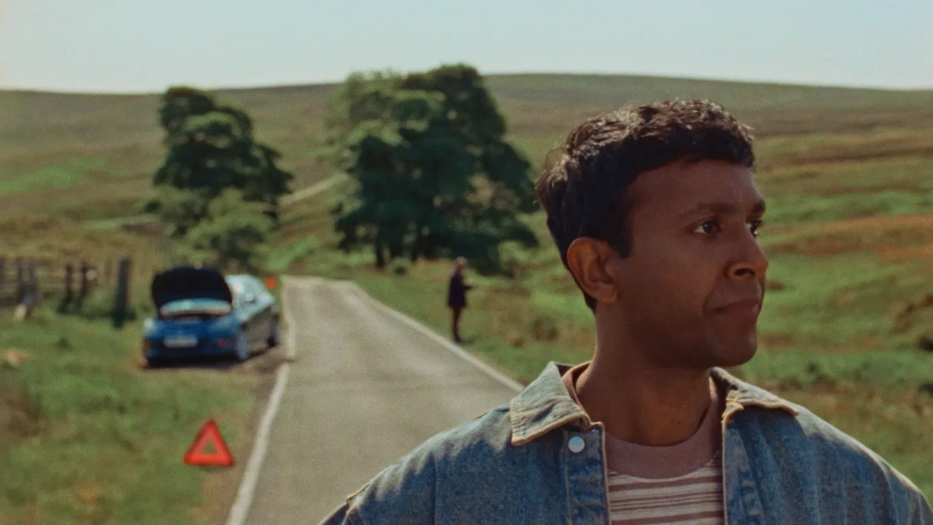 Young man with short dark hair standing on countryside road, roadside car with open trunk, person walking in the distance, green fields, trees, and hills, bright daylight.