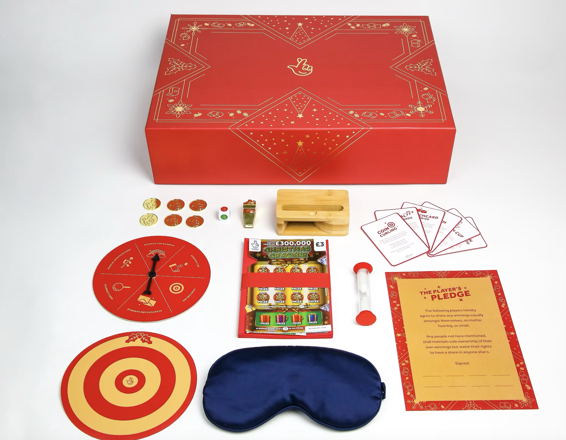 A holiday-themed game set with a red and gold box, a game wheel, poker chips, a small wooden holder, a pack of scratch-off tickets, a paper pledge, a magnifying glass, a blue sleep mask, and other game components arranged on a white surface.