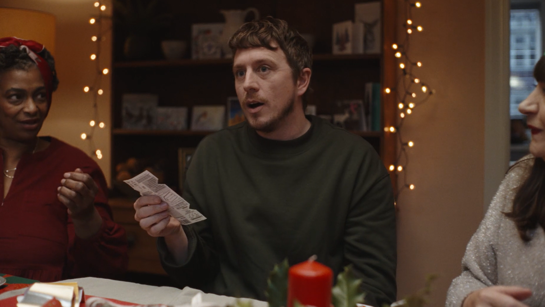 Three people are gathered around a table during a holiday celebration. The woman on the left, wearing a red top and a headband, is looking towards the man in the center. The man is holding a piece of paper and appears to be speaking or explaining. The woman on the right, dressed in a glittery top, listens attentively. The background shows a shelf with framed photos and a string of warm fairy lights, indicating a cozy indoor setting.