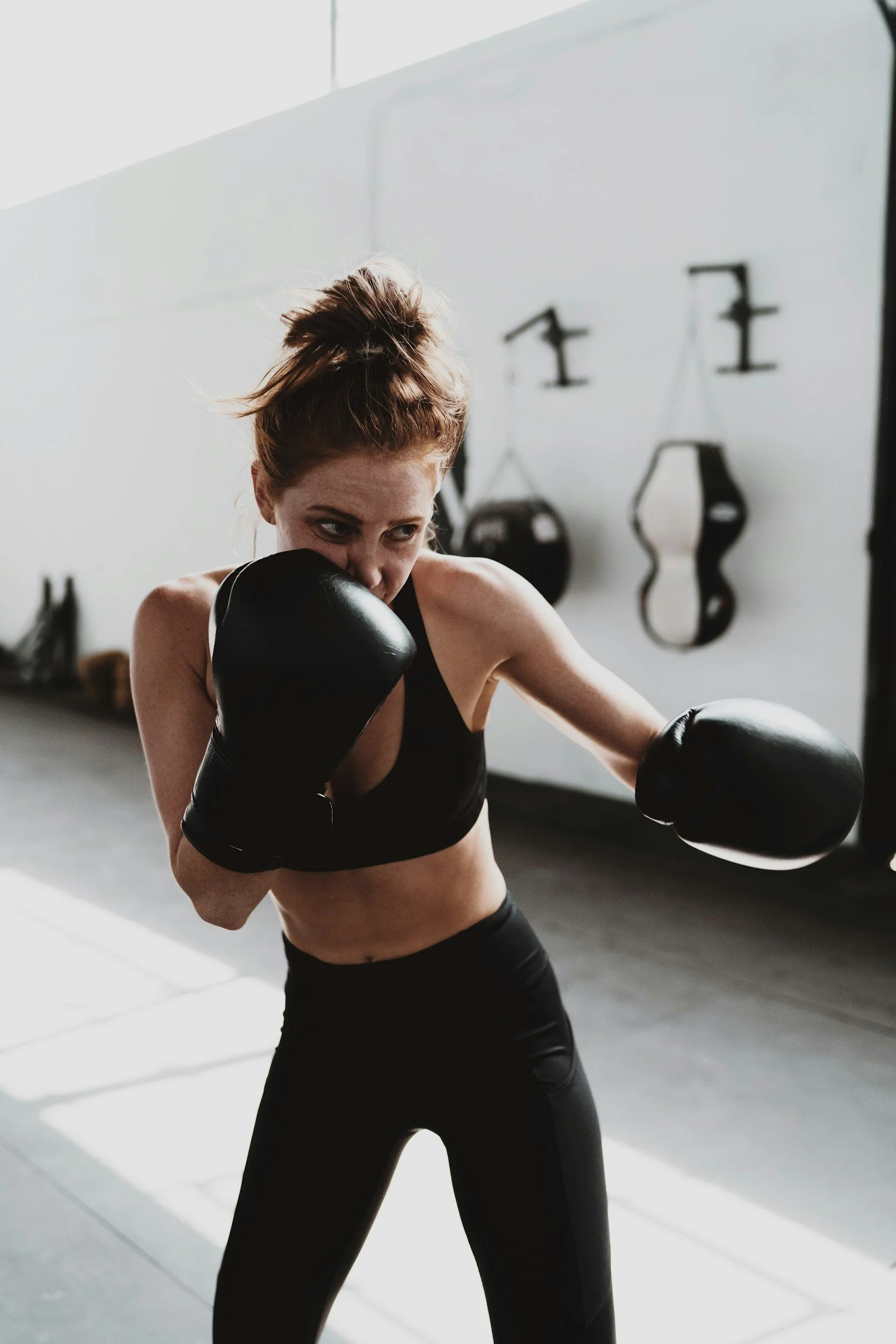 A woman with red hair tied up, dressed in black sportswear, is practicing boxing in a gym, wearing black boxing gloves, with a focused expression.