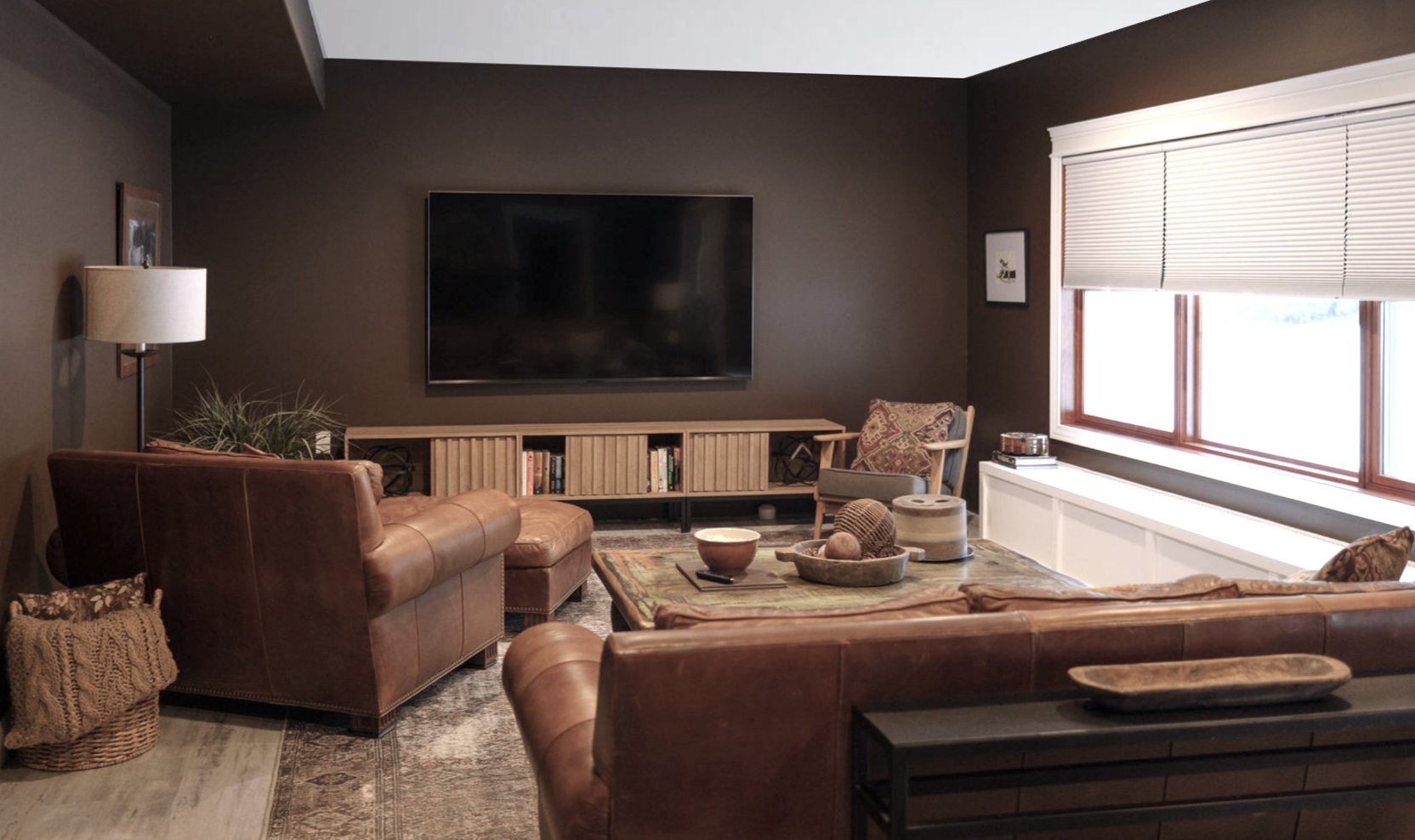 Basement media room with leather sofa and chair. A window seat adds extra storage and seating space. Deep rich brown walls add a cozy feature to the media room and help make a large 85 inch TV feel cohesive and blends in.