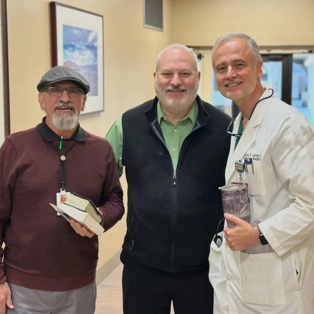 Three smiling men standing together indoors, with the man on the right dressed as a doctor in a white coat, holding a pulse oximeter, and the man on the left holding a book. The man in the center wears a black vest over a green shirt.
