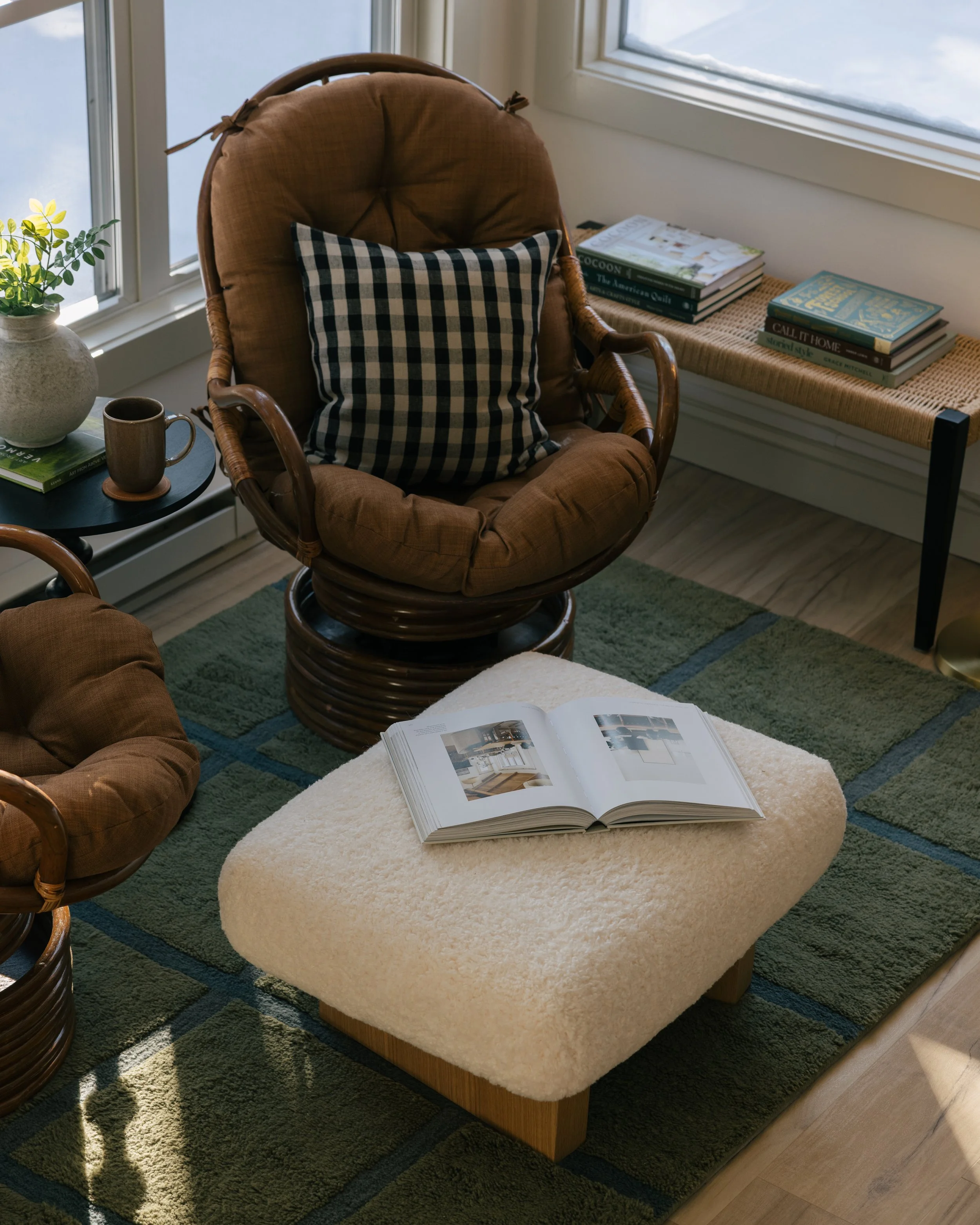 Cozy living room corner with a round rattan armchair with a checkered pillow, a side table with a coffee mug and plant, a cream-colored ottoman with an open book, a green rug, and a window with snowy outdoors visible.